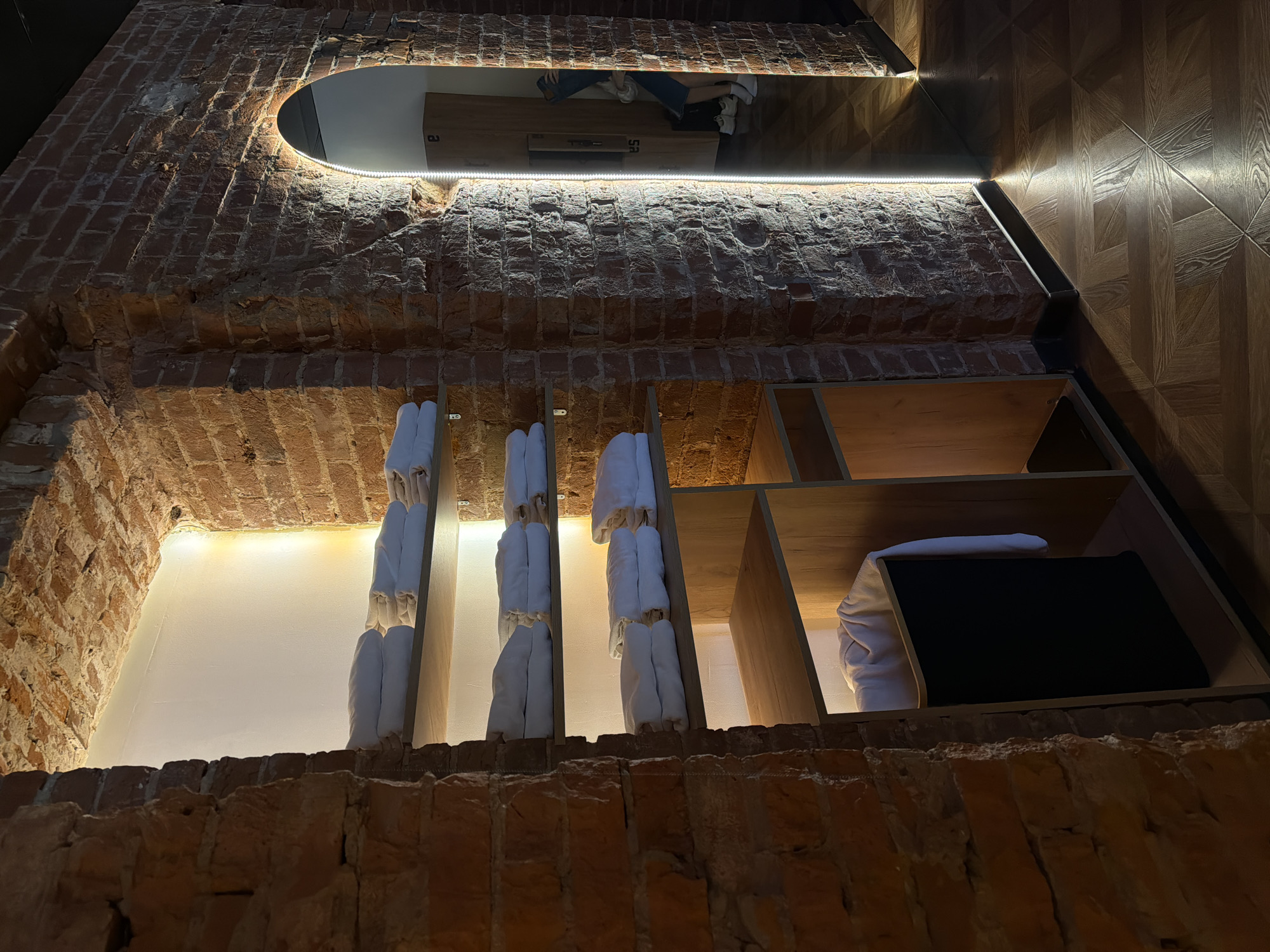 The image presents an unusual perspective, looking upwards at a recessed alcove within a brick wall, possibly an interior design element. The alcove features a mirrored section at the top, illuminated by an integrated light strip. The mirror reflects a scene above and behind the photographer, appearing to show a room with a person. Below the mirror, several white towels are neatly hung or stacked within a wooden shelving unit. To the right of the towels, the shelving continues, containing a rolled-up item and a dark, flat object, perhaps a case or folio. The wall to the right of the alcove is paneled with a decorative wood design. The brickwork of the alcove is prominent, displaying rough textures and varied coloring. Considering the presence of brick and the general aesthetic, it is possibly located in the city of Moscow, Russia.