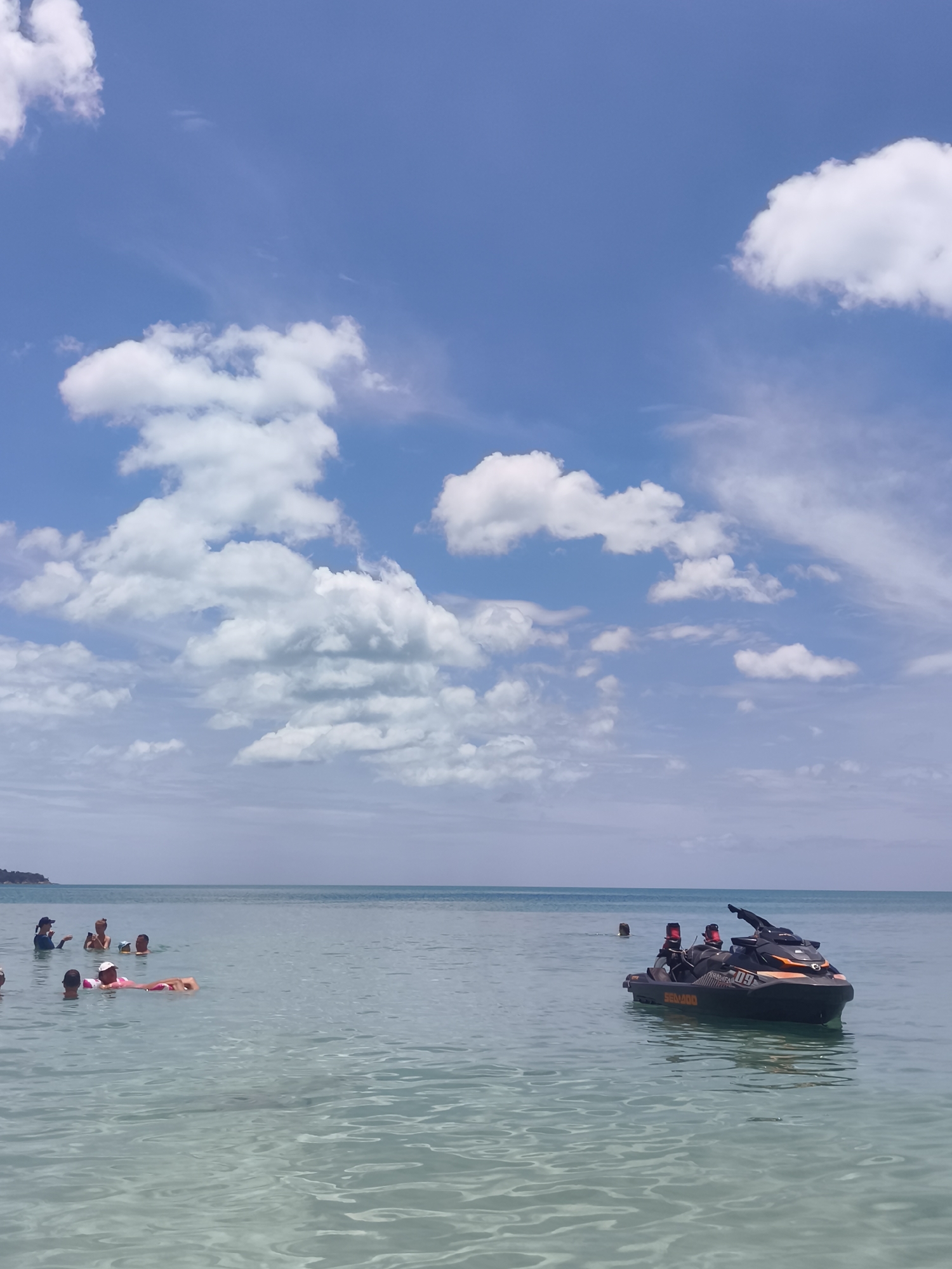In the bright, clear waters of Ko Samui, Thailand, a group of people enjoy a leisurely day. The turquoise sea stretches to the horizon, meeting a sky dotted with fluffy white clouds. In the foreground, several individuals are scattered across the water, some swimming and others relaxing on inflatable floats. A person on a pink float is visible on the left side of the frame.
Further out, a jet ski is being operated by two individuals. They appear to be enjoying a ride on the calm, shallow water. In the background, a distant shoreline is faintly visible, hinting at the lush greenery of the island. The overall scene depicts a serene and inviting tropical paradise.