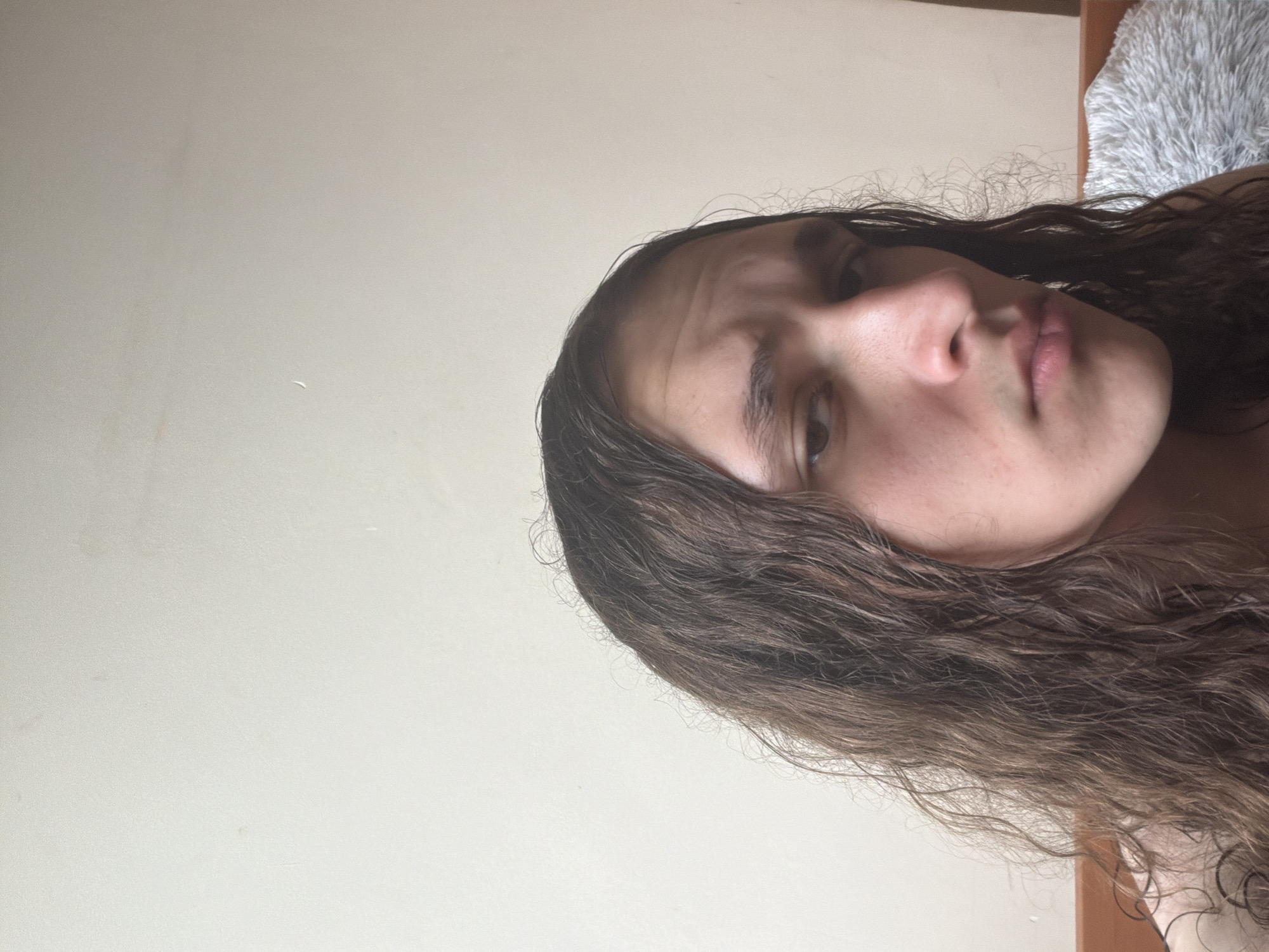 A selfie of a person with long, wavy brown hair is positioned with their head in the lower right corner and their face centered in the frame. The background consists of a plain, light-colored wall. A white fluffy object is partially visible in the upper right corner. The image orientation is horizontal. There is no identifiable evidence to determine that the image was taken in Plovdiv, Bulgaria.