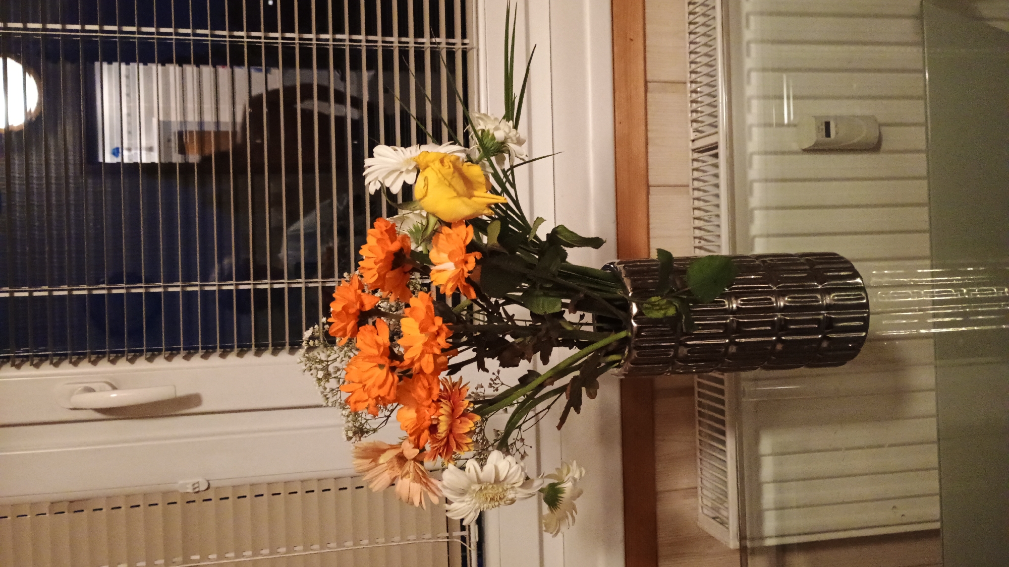 A bouquet of artificial flowers, predominantly orange with some white and a single yellow flower, is placed in a dark, textured vase. The vase sits on a glass surface, reflecting the flowers and vase. Behind the vase, a radiator is visible, and beyond that, a window with blinds. The blinds are partially open, revealing a dark interior with a faint reflection of a person in silhouette, suggesting an indoor environment, likely a residential setting. The overall lighting is dim, with some light coming from the window. The scene appears to be located in Sokolov, Czechia, as indicated by external context. There are no discernible actions or interactions taking place. The description is purely observational and objective.