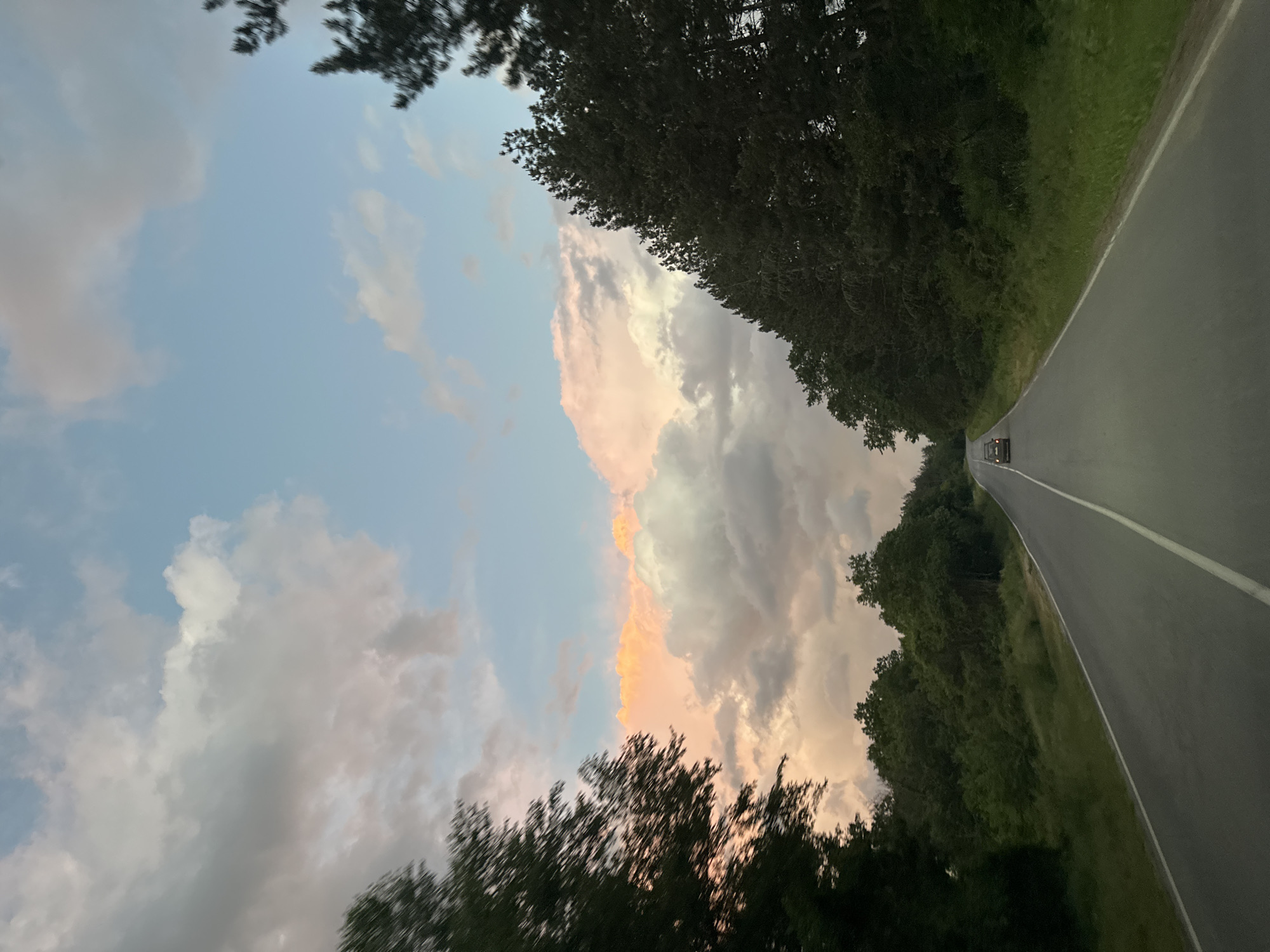 A low-angle, landscape-oriented shot of a road winding through a wooded area. The road, with a painted white line down the middle, curves gently towards the top of the frame. On either side of the road are densely packed trees. The sky is visible above the trees, featuring a mix of blue and grey clouds. There is a hint of pink/orange on some of the clouds, suggesting either a sunrise or sunset. The scene appears to be taken at dusk.
While there are cars and a road, there is insufficient evidence to confirm that the picture was taken near the Bulgarian village of Pryaporets.