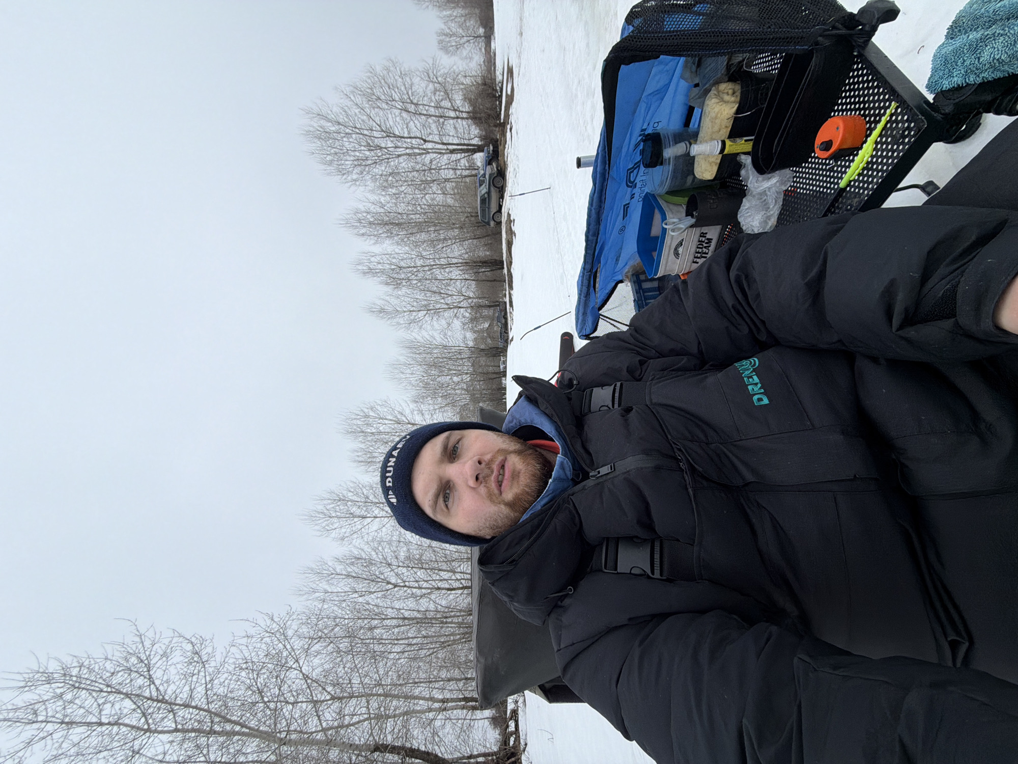 The image depicts a man outdoors in a snowy environment, likely near a body of water. The man is wearing a dark blue beanie, a blue hooded sweatshirt, and a black puffer jacket. To his right is a black and metal fishing station with various fishing supplies, including a blue tarp, containers, and tools. Bare trees are visible in the background, and a vehicle is parked further in the distance. The overcast sky suggests it is winter or a cold day. The precise location within Noviy, Russia, is not discernible from the image.