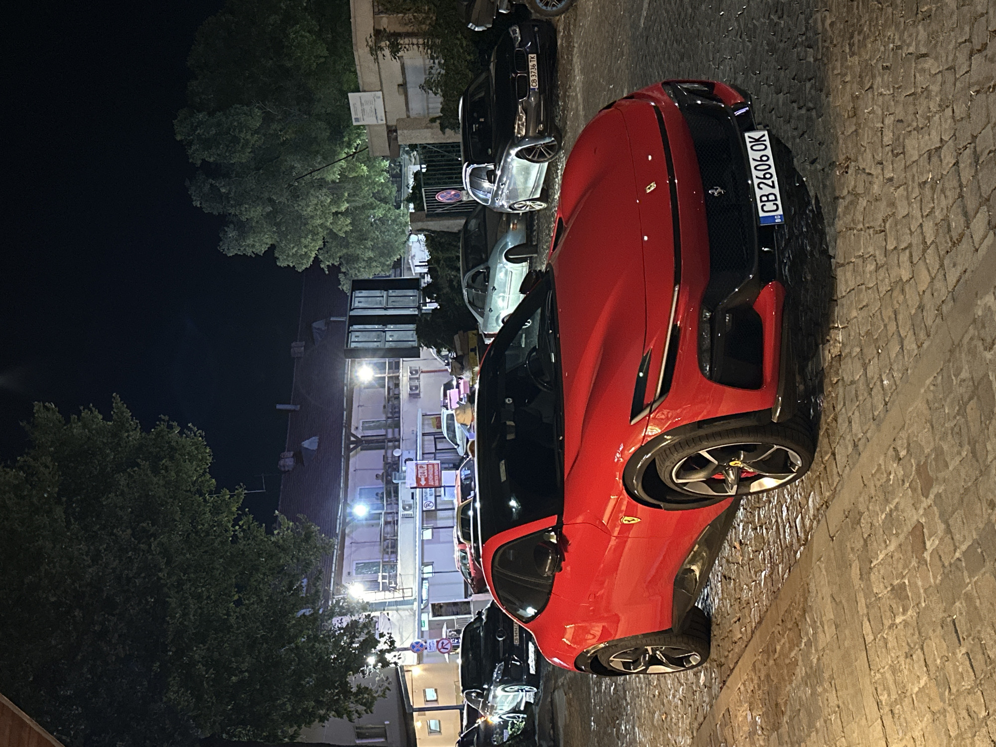 An outdoor, medium-angle, eye-level shot captures a red Ferrari Purosangue parked on a cobblestone street at night in Sozopol, Bulgaria. The car faces towards the viewer with its front prominently displayed. The license plate reads "CB 2606 OK" and has a Bulgarian flag insignia. In the background, several other vehicles are parked along the street, including a black Mercedes-Benz, a silver BMW wagon, and a light blue sedan. Buildings with illuminated windows and balconies line the street, with trees providing a dark silhouette against the night sky. The scene appears to be a public area, possibly a town square or a street in the old town, with architectural elements suggestive of a European setting. The lighting is artificial, creating reflections on the wet cobblestones and car surfaces. No people are clearly visible or interacting with the cars or the environment.