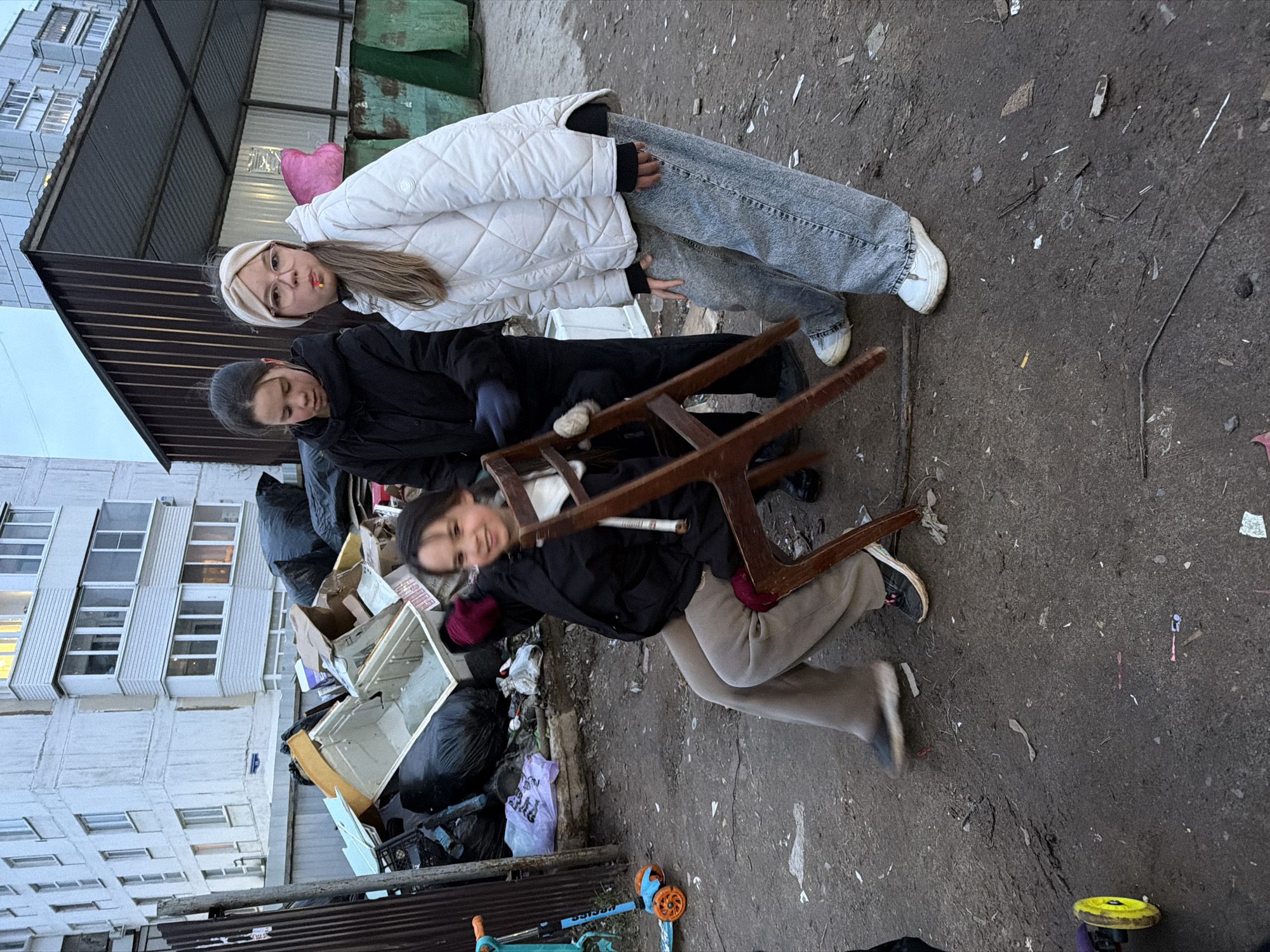 The image captures three young individuals outdoors in what appears to be an urban alleyway or neglected space. The scene is dominated by a dirty, debris-strewn ground and a large pile of trash, including black bags, cardboard boxes, and discarded appliance parts, in the middle ground. In the background, a multi-story, light-colored apartment building with numerous windows rises on the left, while a low metal fence and some green, rusted structures are visible on the right.
Three people are positioned towards the center-right of the frame. The leftmost person, partially obscured, wears a dark jacket and has dark hair. In the center stands a young woman or teenager wearing a white quilted winter jacket, light-wash jeans, and white sneakers. She has long light-brown hair, glasses, and a light-colored headband, looking towards the camera with a slight pout and holding a pink heart-shaped object behind her back. To her right, a younger girl smiles brightly at the camera, dressed in a dark jacket, light brown pants, and dark sneakers. She is playfully holding or swinging a wooden chair, with her hands appearing to be in red gloves or mittens.
The setting suggests a cool or cold day, given the subjects' winter attire. The lighting is soft, indicating either an overcast sky or late afternoon. On the ground near the younger girl, a light blue and orange scooter is visible, along with a yellow disc-shaped object. In the trash pile, a purple plastic bag has some indecipherable text on it. The overall mood conveyed by the subjects is informal and somewhat playful, contrasting with the unkempt surroundings.