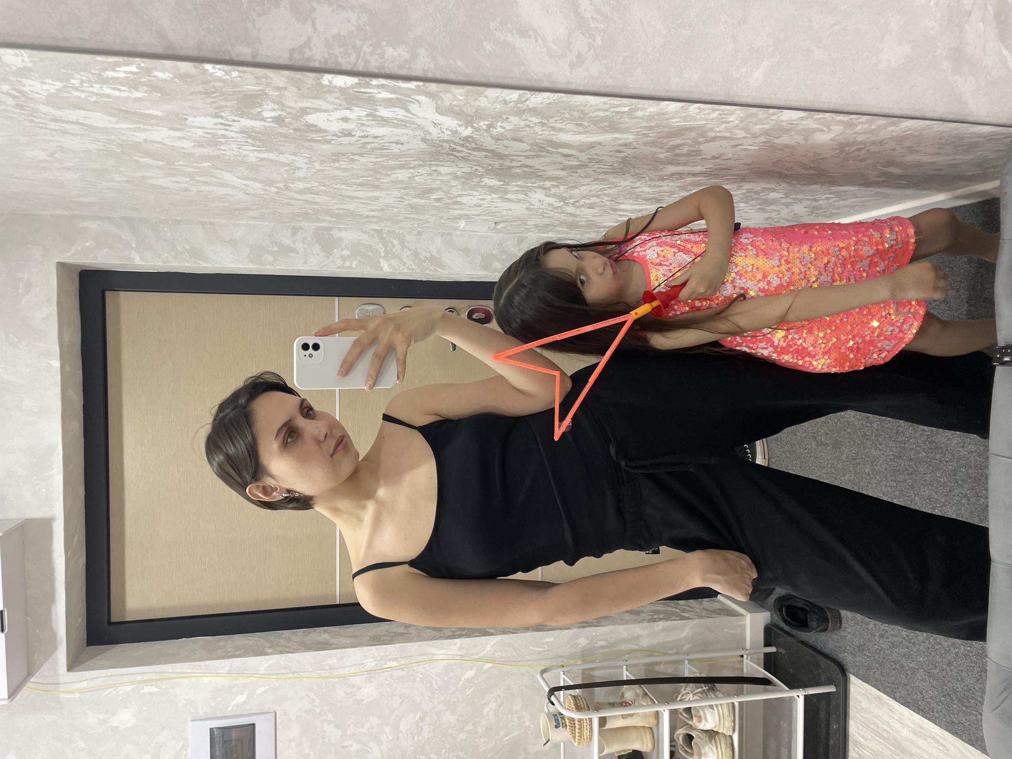The image, taken in Izhevsk, Russia, captures an indoor mirror selfie featuring two females. In the foreground, an adult woman with short dark hair, dressed in a black spaghetti strap top and black wide-leg pants, holds a white smartphone to take the photo. Her reflection in the mirror shows her looking towards the camera with a neutral expression.
Behind and to the right of the adult, a younger girl is visible in the mirror's reflection. She has long dark hair and wears a vibrant, shimmering dress adorned with pink and orange sequins. The girl holds an orange and yellow toy, possibly a bubble wand, and looks towards the camera with a curious or slightly serious expression.
The setting appears to be a room with light-colored, textured walls, possibly wallpapered, visible in the mirror's reflection. The mirror itself has a prominent black frame. The flooring in the room varies; a dark gray textured surface is seen in the lower left, while a lighter gray, possibly carpeted, area is visible on the right. In the bottom right corner, a white multi-tiered shoe rack holds several pairs of light-colored sneakers and shoes. A white rectangular light switch or outlet cover is mounted on the wall to the left of the adult, and a white box is on a shelf further left. A faint yellow line runs along the base of the wall. Subtle text, possibly a "0" or "O", is visible on the adult's black pants. The room is brightly lit by indoor lighting.