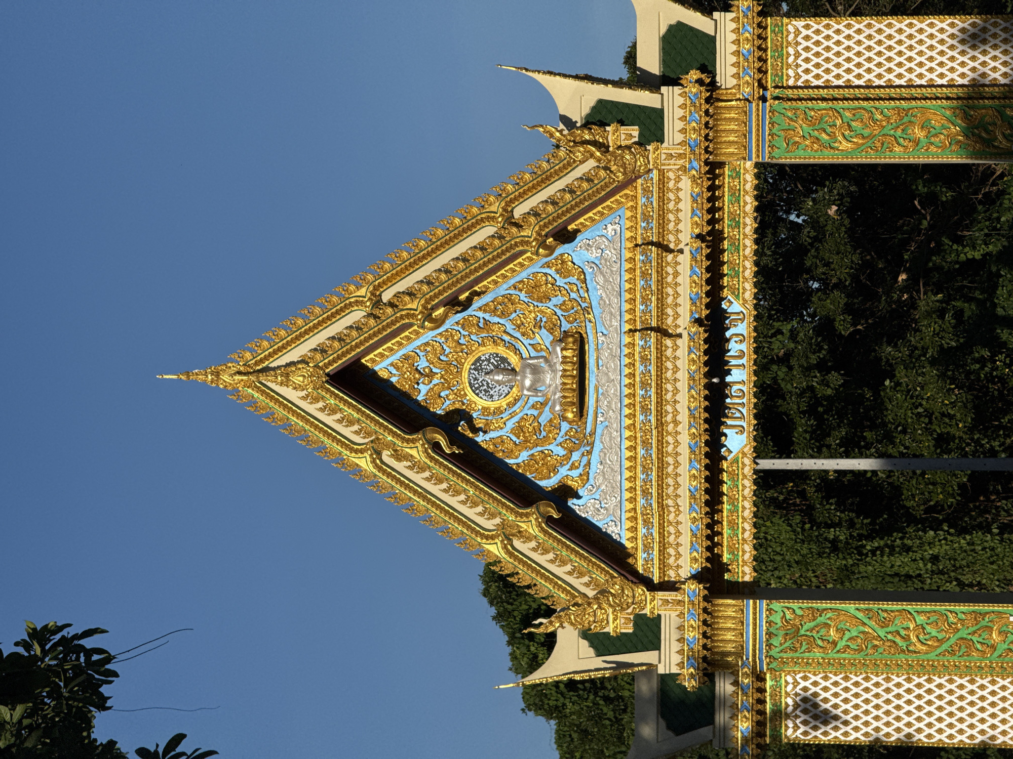 This image depicts the ornate entrance to a temple in Baan Tha Po, Thailand. The entrance features a steep, triangular roof adorned with intricate golden decorations and blue accents. At the apex of the roof, a silver Buddha image sits within a circular frame surrounded by golden floral patterns. The entrance is supported by two pillars, also richly decorated with gold and green ornamental designs. The background is a clear blue sky and lush green trees. The overall impression is one of elaborate craftsmanship and religious significance.