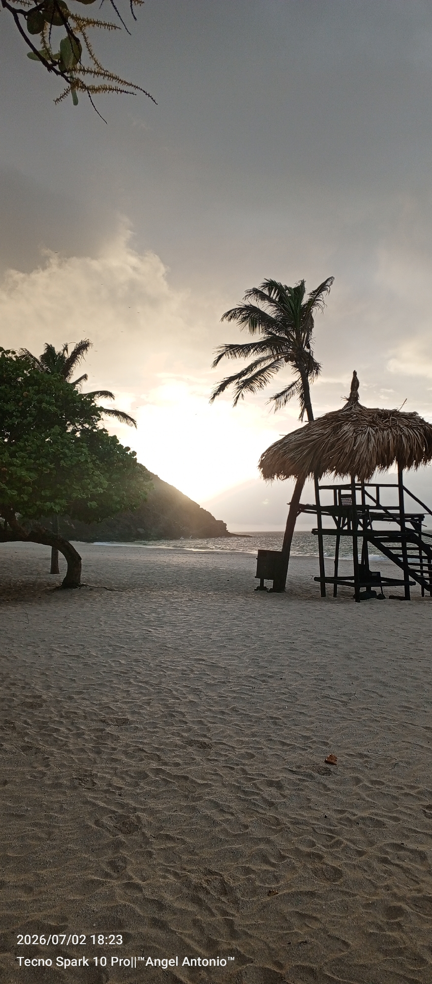 This is a photograph taken on July 2, 2026, at 6:23 PM. The scene depicts a beach in Pedro González, Venezuela, during what appears to be sunset or a cloudy dusk. In the foreground, a sandy beach stretches out, marked by ripples from the receding tide or wind. To the left, a lush green tree with a curved trunk is situated near the water's edge. To the right, a tall, slender palm tree stands beside a rustic, elevated structure with a thatched roof, resembling a lifeguard station or observation post. The structure has wooden stairs leading up to it, and a dark bin is placed next to its base. The ocean is visible in the mid-ground, with gentle waves lapping against a distant, verdant hill that is partially illuminated by the fading sunlight. The sky is overcast with dramatic clouds, and the setting sun casts a soft, diffused light across the scene, creating a serene and tranquil atmosphere. The image is watermarked with "Tecno Spark 10 Pro||™Angel Antonio™".