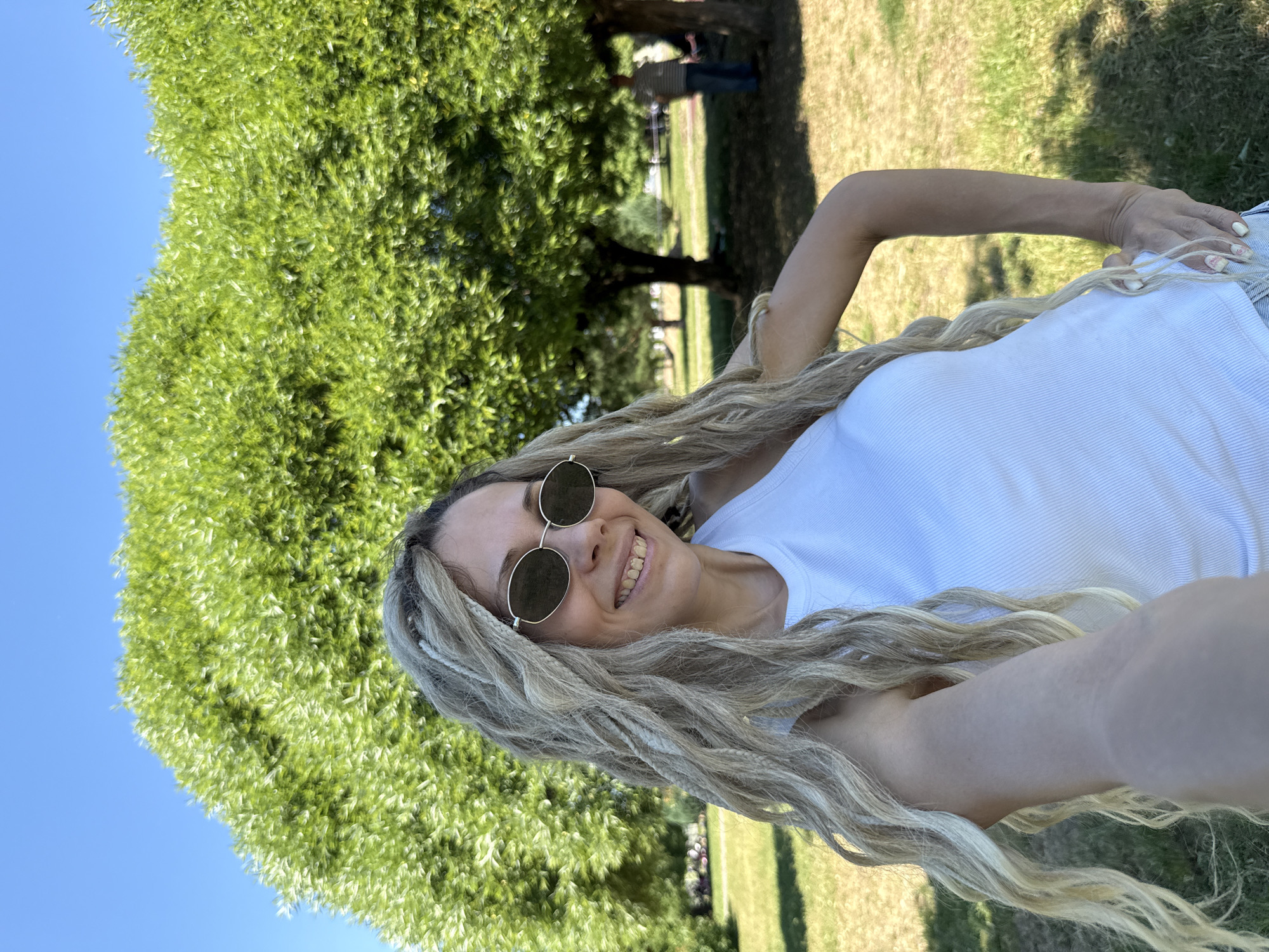 In a sun-drenched park in Zavolzhye, Russia, a woman with long blonde dreadlocks and sunglasses poses for a selfie. She is wearing a white tank top and her right arm is bent at the elbow, with her hand resting on her hip. Behind her, a large, lush green tree dominates the frame, its branches reaching towards a clear blue sky. The park setting is evident with grass and other trees visible in the background, along with a few other people in the distance. The overall impression is one of a bright, warm day in an outdoor recreational area.