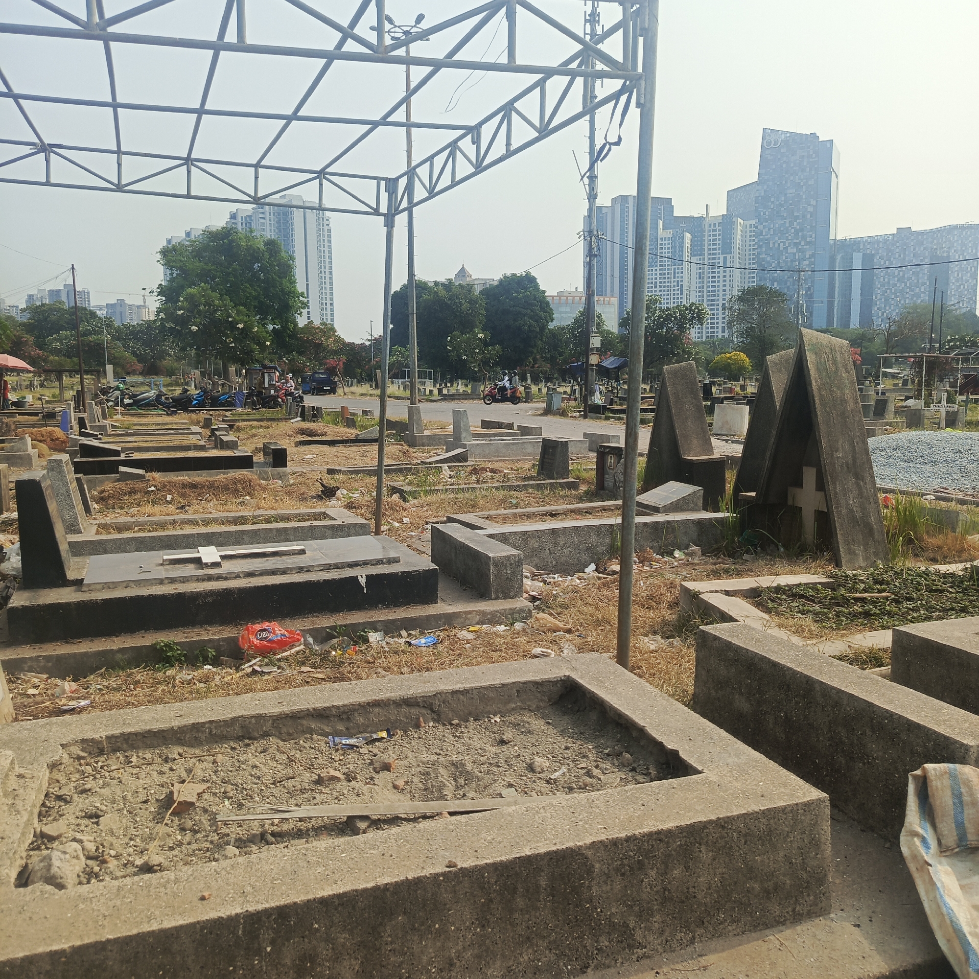 This image depicts a cemetery in South Jakarta, Indonesia, characterized by rows of tombstones and grave sites. In the foreground, several concrete grave borders are visible, some filled with dirt and debris, while others are empty. A prominent metal scaffolding structure dominates the upper portion of the image, suggesting ongoing construction or maintenance work. In the background, modern high-rise buildings rise above trees, indicating the cemetery's urban setting. A road runs through the cemetery with a few motorcycles and a vehicle present, implying some level of activity or passage. Scattered litter is noticeable around the grave sites. The sky is overcast, contributing to the muted lighting of the scene.