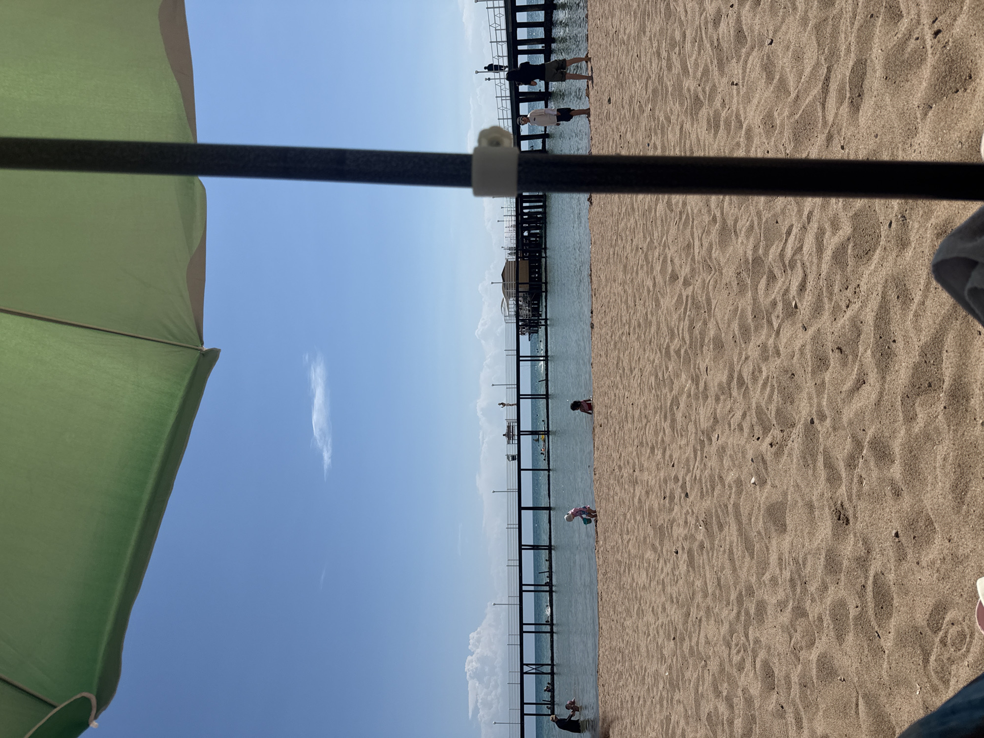 An eye-level, medium shot captures a sun-drenched beach scene at Issyk-Kul Lake, Sary-Oy, Kyrgyzstan. The foreground is dominated by rippled sand, with the dark, vertical pole of a partially visible umbrella casting a shadow. Beyond the sand, the calm, blue water of the lake stretches towards a long, wooden pier supported by numerous posts. People are scattered across the scene: children play in the shallow water near the shore, while adults and children walk along the pier, some sitting on benches. A small, wooden structure with a roof is visible on the pier. The sky is a clear, vibrant blue with a few wispy white clouds. The overall impression is one of a peaceful, sunny day at a popular lakeside resort.