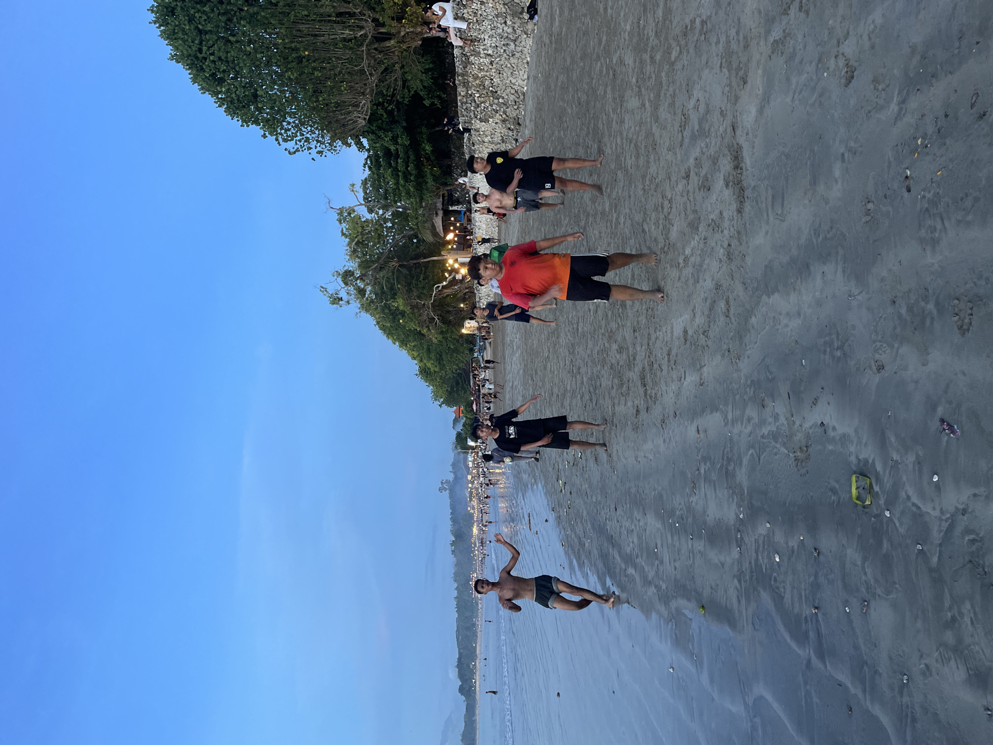 The image depicts a lively beach scene during what appears to be dusk or early evening, likely in Kuta Selatan, Indonesia. The sky above is a clear, soft blue, transitioning into a lighter hue towards the horizon.
In the foreground and midground, several young men are walking or standing on the sandy shore. One man in the foreground, wearing dark shorts and no shirt, appears to be in motion, perhaps mid-jump or posing. Further back, another man in a vibrant orange and black t-shirt and dark shorts stands barefoot on the sand, looking towards the camera. Behind him, a man in a black t-shirt with a yellow emblem (possibly the number "8") and black shorts stands with his arms outstretched. Another shirtless man in dark shorts is visible behind them. All individuals are barefoot.
The beach itself consists of a mix of light and dark sand, with parts appearing wet and reflective, mirroring the sky. Small stones and shell fragments are scattered across the drier areas of the sand. To the left, the edge of the water is visible, showing gentle waves or the tideline.
Along the upper edge of the beach, a dense line of tropical trees and foliage creates a natural barrier. Beneath the trees, numerous artificial lights, possibly from restaurants or stalls, illuminate a bustling area where more people can be seen gathered and walking. The overall atmosphere is casual and relaxed, characteristic of a popular beach destination at the end of the day. A distinct yellow object, possibly a plastic container, is visible on the sand in the mid-right.