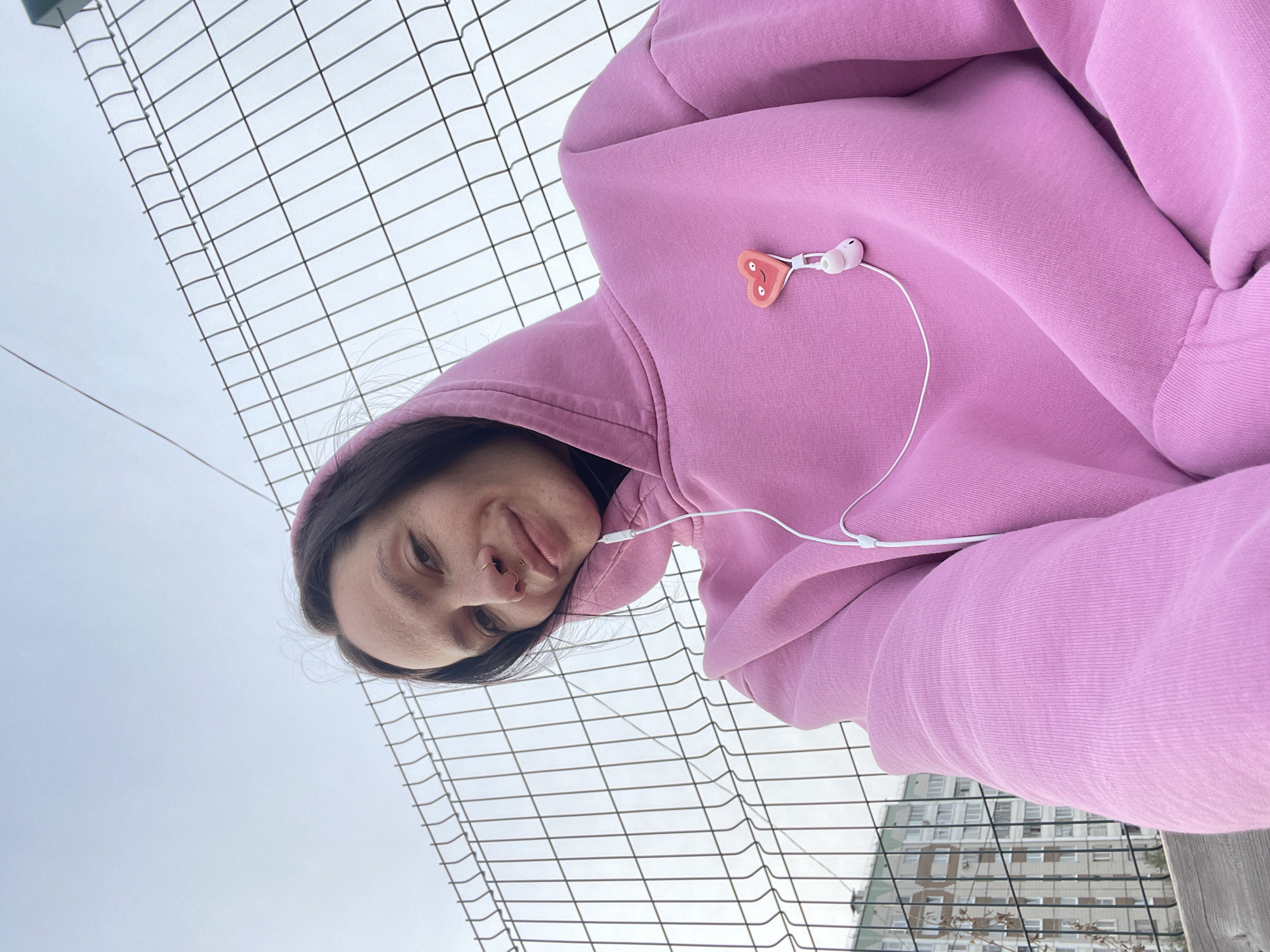 A woman with dark hair is wearing a pink hoodie and white headphones while taking a selfie against a grey, cloudy sky and a metal chain-link fence. A building is visible behind the fence. Based on visual characteristics of the buildings present in the background, this image can be geolocated to the city of Izhevsk, Russia.