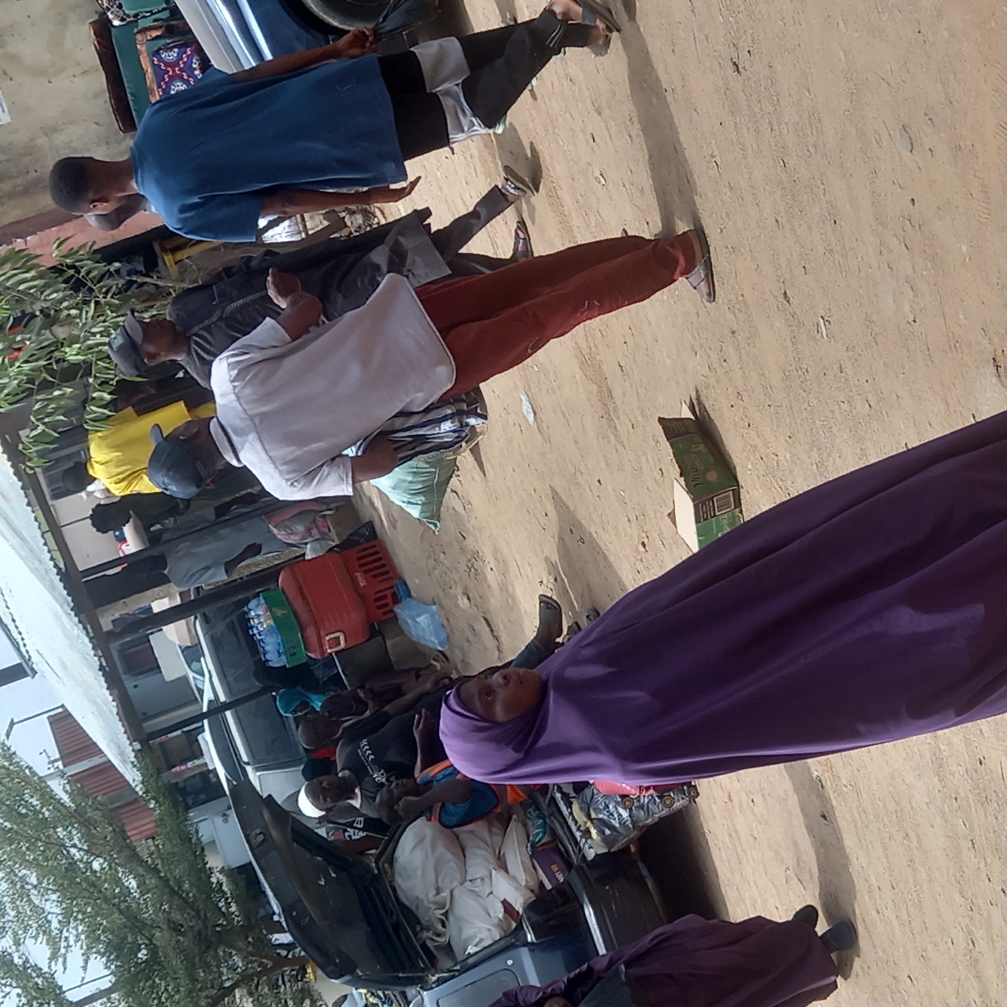 The image shows a group of people in Dadi Riba, Nigeria, likely at a roadside or market area. Several individuals are visible, including a woman wearing a purple hijab. There are men wearing casual clothing, such as blue t-shirts, a white shirt, and rust-colored pants. A car is present, loaded with goods and luggage. The environment appears to be a dusty, outdoor setting. There is a building or structure in the background. Some individuals are standing, while others seem to be seated near the car. There are boxes and various items loaded onto the vehicle, suggesting they are either setting up a roadside market, traveling, or recently arrived.