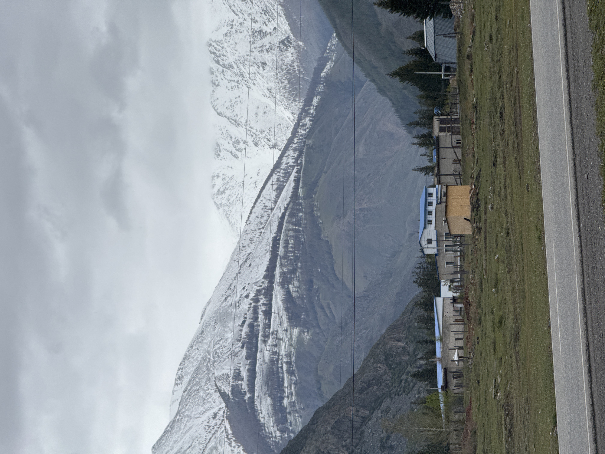 The image shows a mountain landscape near Aktash, Russia. A large mountain partially covered in snow dominates the background, disappearing into a cloudy sky. A cluster of buildings with blue and brown roofs stands along a green, grassy area beside a gray paved road on the right side of the image. Power lines are visible in the foreground, stretching across the mountain.