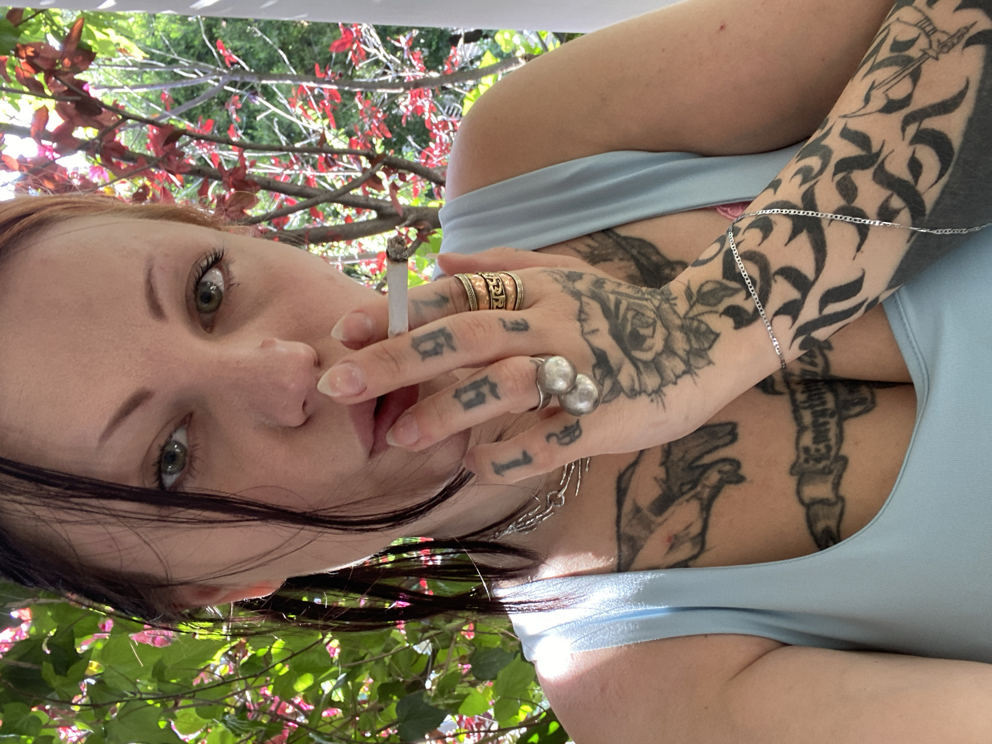 The image depicts a woman with fair skin, dark hair, and green eyes. She is holding a lit cigarette to her lips. The woman has numerous tattoos covering her arms, chest, and hands, including script, floral designs, and what appears to be a bird. She wears multiple rings on her fingers, including a large, ornate gold ring and a silver ring with two spheres. She is wearing a light blue, low-cut top. The background features foliage with red and green leaves. There is no information to ascertain the location is in Kunje, Montenegro.