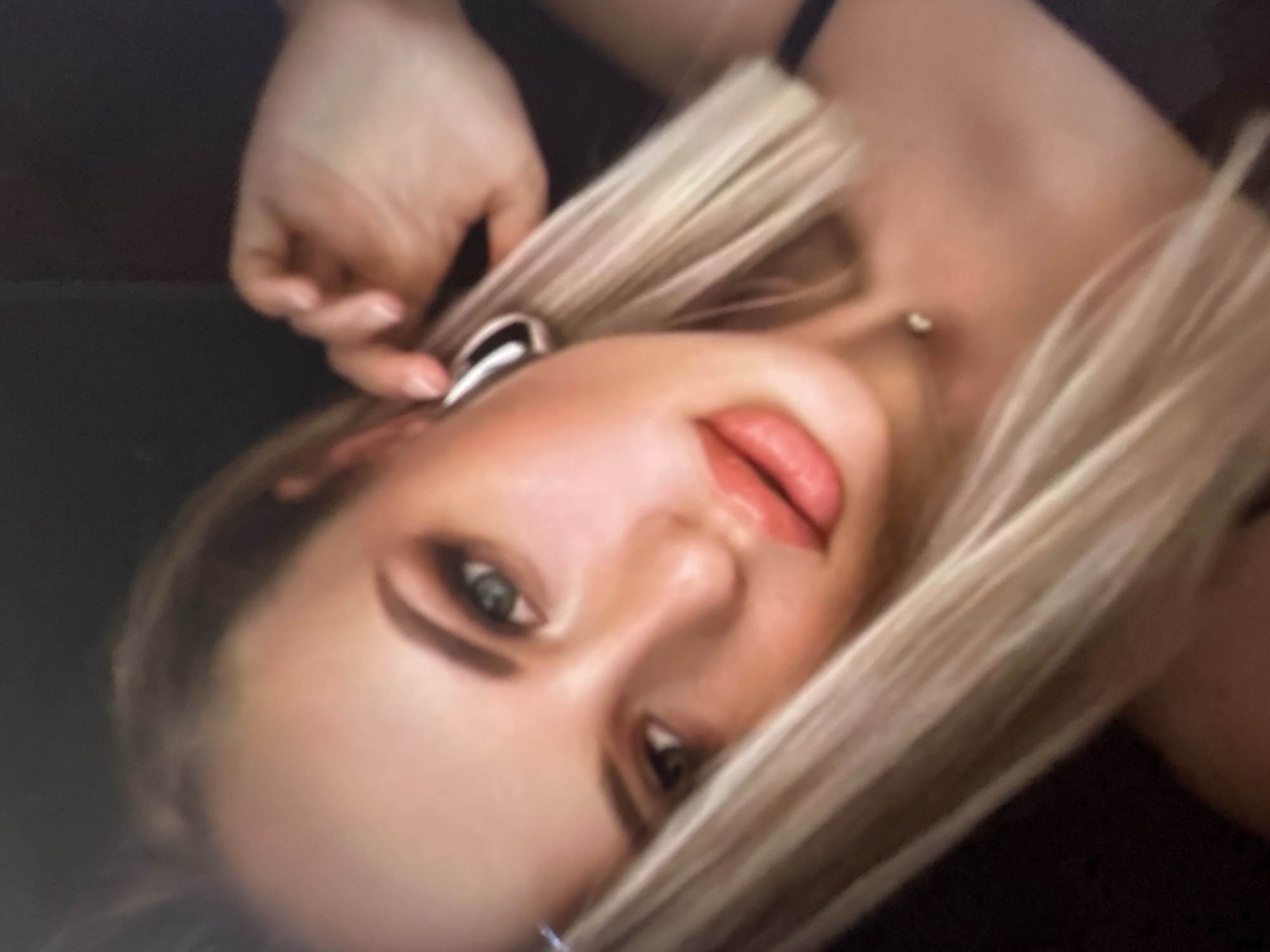 The image shows a close-up, blurry shot of a young woman with blonde hair and fair skin. She is wearing makeup, including eyeshadow and lipstick. She has large hoop earrings and a small silver necklace with a pendant. Her right hand is touching her ear, and her left hand is not visible. The background is dark and out of focus. The image quality is low, and the subject is the only discernible element. There is no information in the image that allows for the determination of its location in Gelendzhik, Russia.