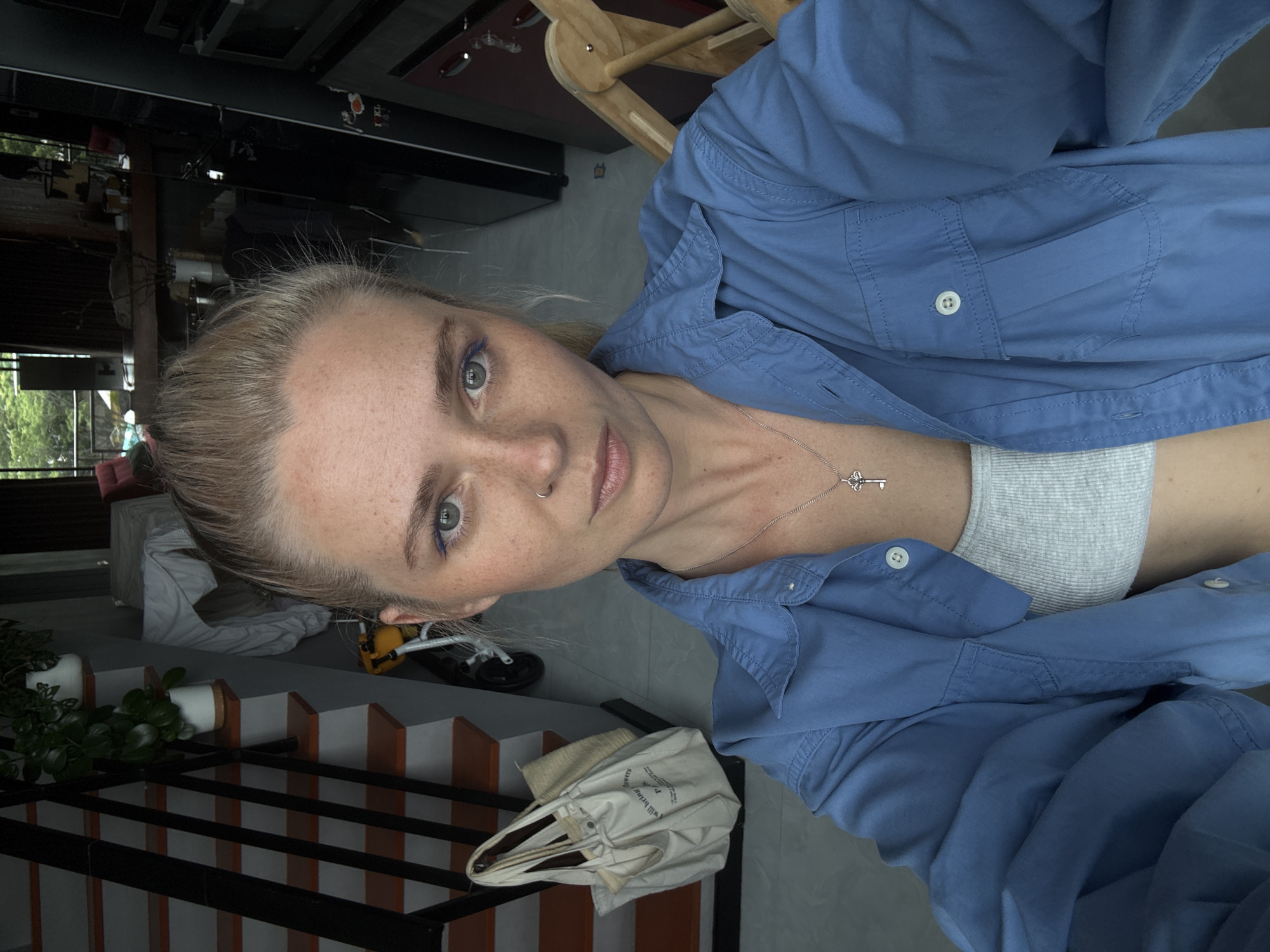 A selfie taken from the chest up shows a young woman with blonde hair pulled back in a ponytail, freckles, and light blue eyeliner. She is wearing a light blue collared shirt unbuttoned over a gray ribbed tank top and a silver necklace with a key pendant. The background appears to be an interior space, possibly a home, with a staircase on the left, a kitchen area with appliances on the right, and an outdoor view with greenery visible through windows. The scene is lit with natural light.