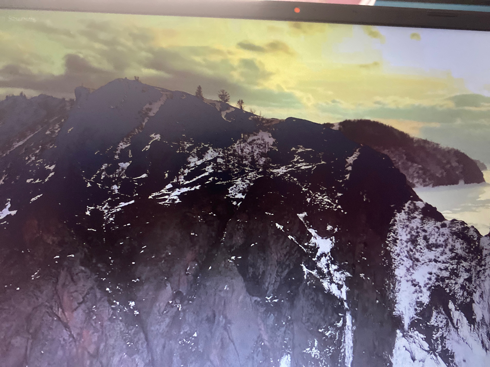 The image displays a dramatic mountainous landscape under a cloudy, yellow-tinged sky. The mountains are dark and rugged, with patches of snow scattered across their slopes, particularly on the foreground mountain. Some sparse trees are visible on the higher ridges. In the distance, another mountain range is partially obscured by mist or clouds. The overall impression is one of a remote and somewhat desolate natural environment. The location is not specified as being in Hemayetpur, Bangladesh.