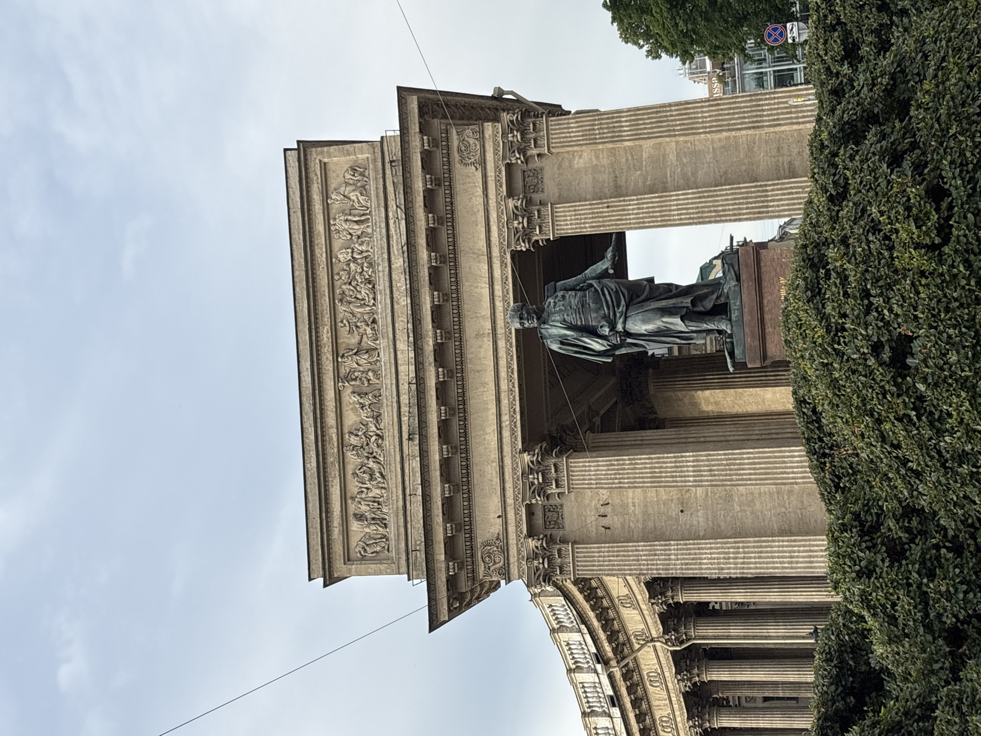 This image captures the Kazan Cathedral in Saint Petersburg, Russia, a prominent landmark on Nevsky Prospekt. In the foreground, a dense green hedge obscures the lower part of the scene. Rising above the hedge is a majestic bronze statue of Field Marshal Mikhail Kutuzov, depicted in military attire with a cloak draped over his shoulders and his left hand extended. Behind the statue, the imposing colonnade of the Kazan Cathedral dominates the frame. The facade is characterized by fluted Corinthian columns and a pediment adorned with bas-relief sculptures depicting classical figures. The sky above is overcast, with faint clouds and the thin lines of overhead wires crossing the frame. To the right, a glimpse of modern city buildings and a traffic sign can be seen, contrasting with the historical architecture.