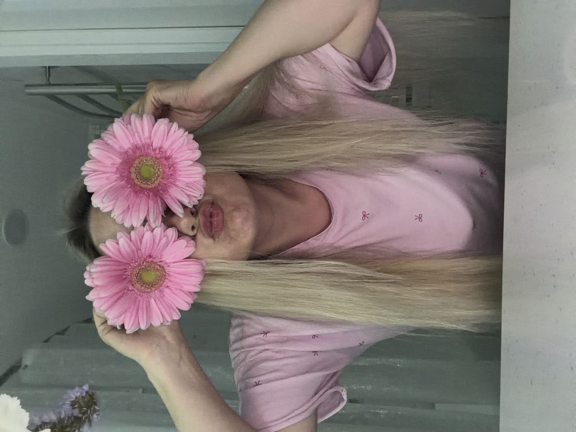 A light-skinned individual with blond hair is holding two pink gerbera daisies in front of their face. They are wearing a light pink shirt with a bow pattern. The background appears to be a bathroom with a shower fixture visible. There is no indication that this image was taken in Votkinsk, Russia.