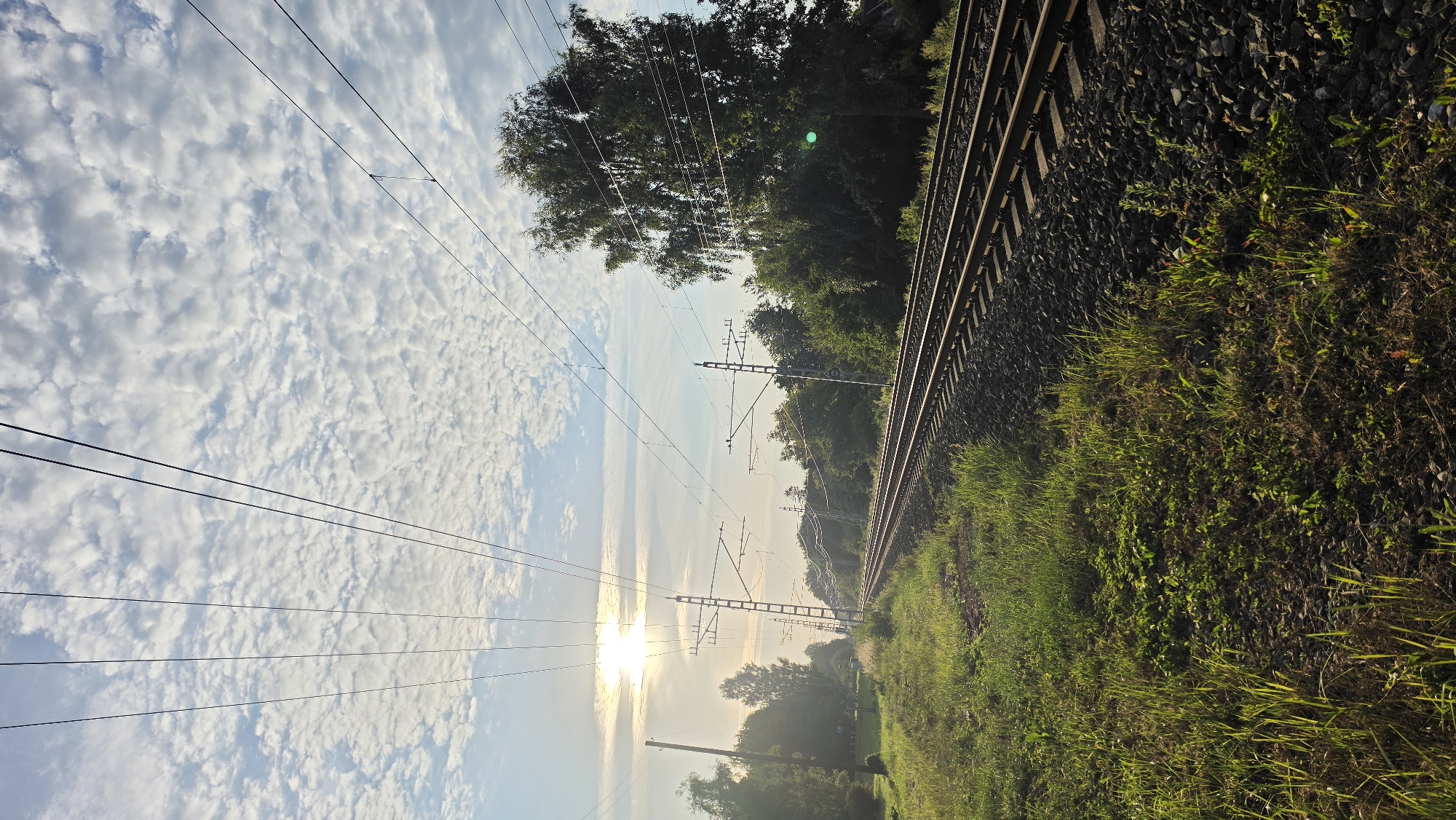 The image shows a railroad track in a rural or suburban area. The sun is visible in the sky, casting a warm glow on the scene. There are trees and grass on either side of the tracks, and power lines run overhead. The sky is partly cloudy, with a textured pattern of small clouds. The overall impression is one of a peaceful, natural setting. The image is taken from a low angle, looking down the tracks. The lighting suggests it is either early morning or late afternoon. The railroad tracks appear to be electrified, as indicated by the presence of overhead power lines and supporting structures. The vegetation along the tracks is lush and green, suggesting a temperate climate. The ground around the tracks is covered in ballast, which is typical for railway construction. The image does not contain any people or any specific landmarks that would identify its exact location within Jūrmala, Latvia.