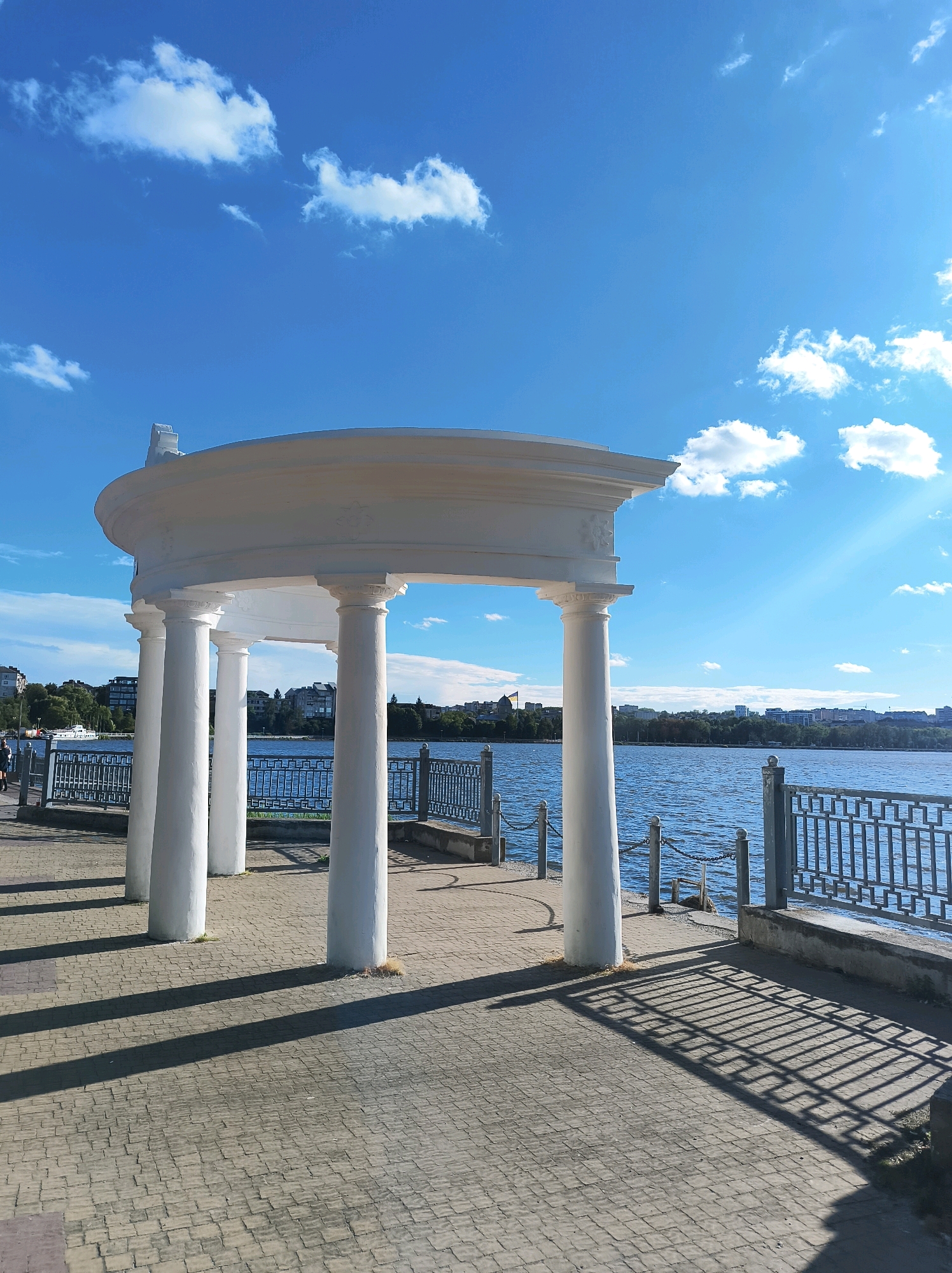 The image depicts a waterfront promenade in Ternopil, Ukraine, featuring a white, classical-style gazebo with Corinthian columns. The gazebo overlooks a calm blue body of water, likely Ternopil Lake, with a distant cityscape visible on the opposite shore. A metal railing lines the waterfront. The ground is paved with interlocking grey bricks, casting long shadows from the gazebo's columns. The sky is a vibrant blue with scattered white clouds, and the sun is shining, indicated by the strong shadows and light rays. A white boat is docked on the water, and a few people are walking along the promenade in the background to the left. The overall scene conveys a peaceful, sunny day at a popular urban waterfront.