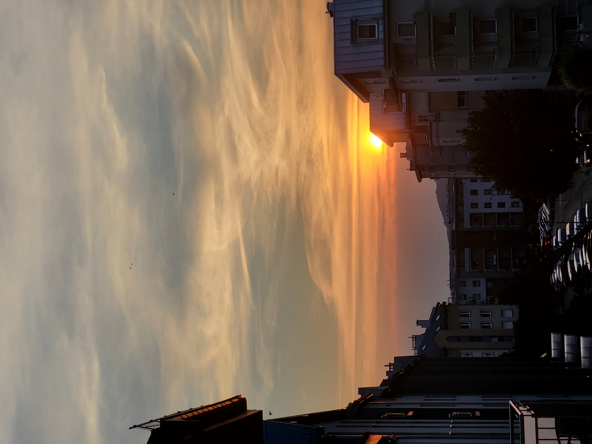The image shows a sunset over a city, with buildings and a sky filled with clouds. The sun is low on the horizon, casting a warm glow. There are several buildings visible, with a mix of architectural styles. Some of the buildings appear to be apartment complexes, while others are smaller. The sky is a vibrant orange and yellow, with streaks of white and gray clouds. There are a few birds visible in the sky. The overall impression is one of a peaceful evening in an urban environment.