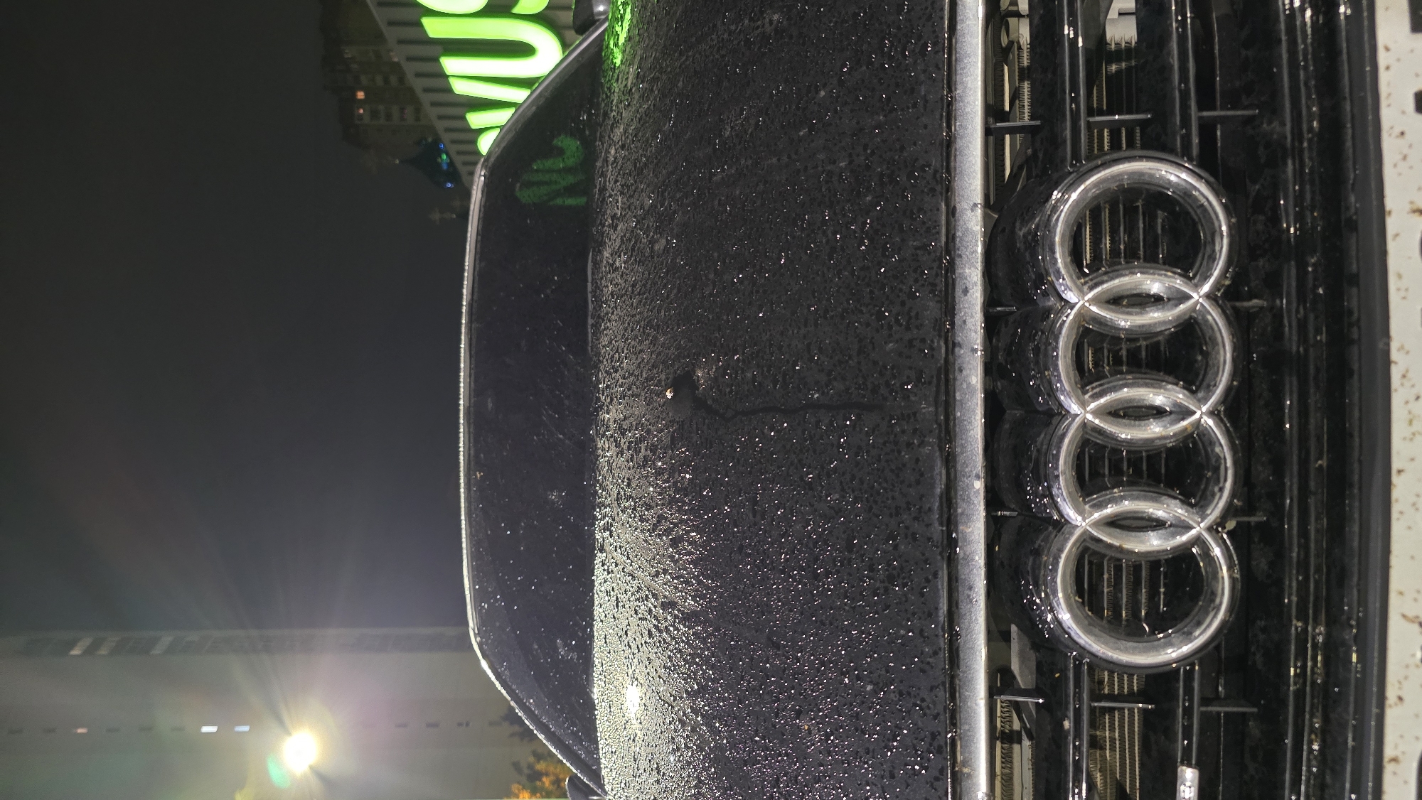 A close-up photograph taken at night depicts the front of a dark-colored Audi car. The car's hood, leading to the front grille, is heavily speckled with numerous small water droplets, indicating recent rain or condensation. The prominent Audi four-ring logo is centered on the black grille, which features a vertical slat design. A single thin streak of water runs down the wet hood from a brighter droplet. In the background, against a dark night sky, bright artificial lights cast lens flares, particularly in the lower left. A large, green illuminated sign is partially visible in the upper right background, displaying fragmented letters such as "VUS" or similar. Faint building structures are also discernible in the distance. The scene suggests an urban or suburban environment at night, after rainfall. No specific activity is taking place; it's a static shot of the vehicle.