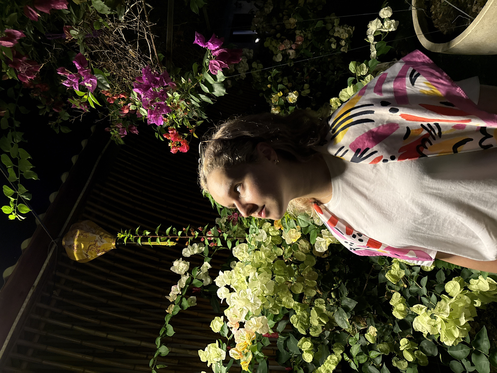The image captures a young woman in Phú Quốc, Vietnam, seemingly at night. She has light skin, curly blond hair, and is wearing a white t-shirt. A patterned scarf or wrap is draped over her shoulder. The woman is surrounded by greenery, with a variety of flowering plants in shades of pink, red, and white. A decorative hanging light fixture is visible against a background that appears to be a building with a bamboo or similarly textured facade.