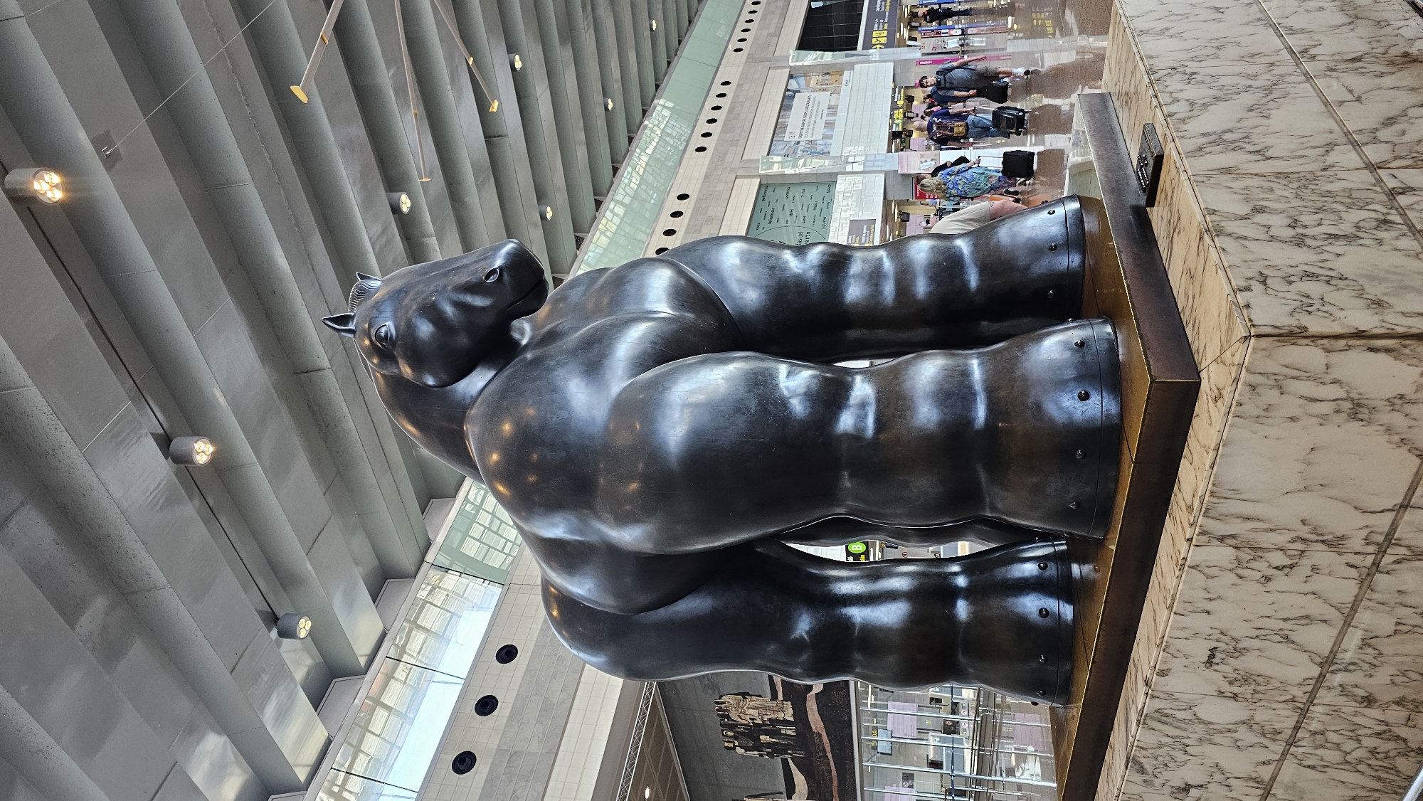 The image shows Fernando Botero's "Horse with Bridle", a large, bronze sculpture of a horse, located at Terminal 1 of Barcelona El Prat Airport, in El Prat de Llobregat, Spain. The sculpture is prominently featured in the image. The statue is set upon a wooden pedestal. The airport's architecture is visible in the background, including a ribbed ceiling with light fixtures, and glass walls. People are seen in the blurred background, suggesting an active airport environment. The floor is marble.