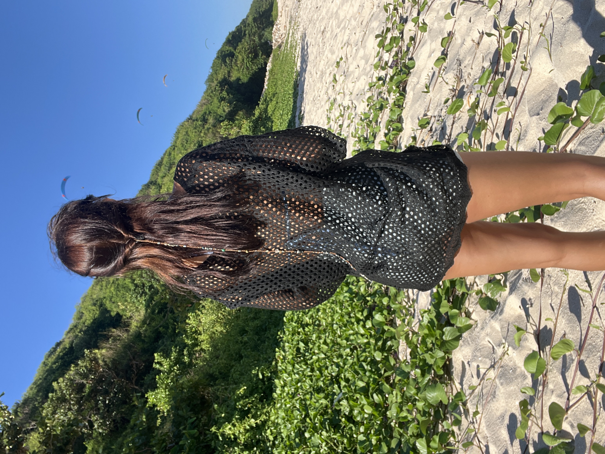 The image displays a person standing on a sandy path, viewed from behind, against a backdrop of lush greenery and a clear blue sky. The person has long, dark brown hair, which appears to be styled or partially pulled back, revealing some delicate beads or a chain accessory intertwined within it. They are wearing a black, sheer, mesh-patterned cover-up that extends down to their upper thighs, showcasing tanned skin underneath.
The ground consists of light-colored sand interspersed with vibrant green creeping or ground-cover plants. In the mid-ground and background, a large, verdant hill or cliff covered with dense tropical foliage dominates the left side of the frame. Above the hill, the sky is a brilliant, cloudless blue. Scattered across the vast expanse of the sky are several paragliders, appearing as small, colorful arcs. The bright sunlight suggests it is daytime with clear, sunny weather, casting distinct shadows on the sandy ground. The scene evokes a sense of outdoor leisure and natural beauty, characteristic of a coastal or hilly location such as Kuta Selatan, Indonesia. No visible text is present in the image.