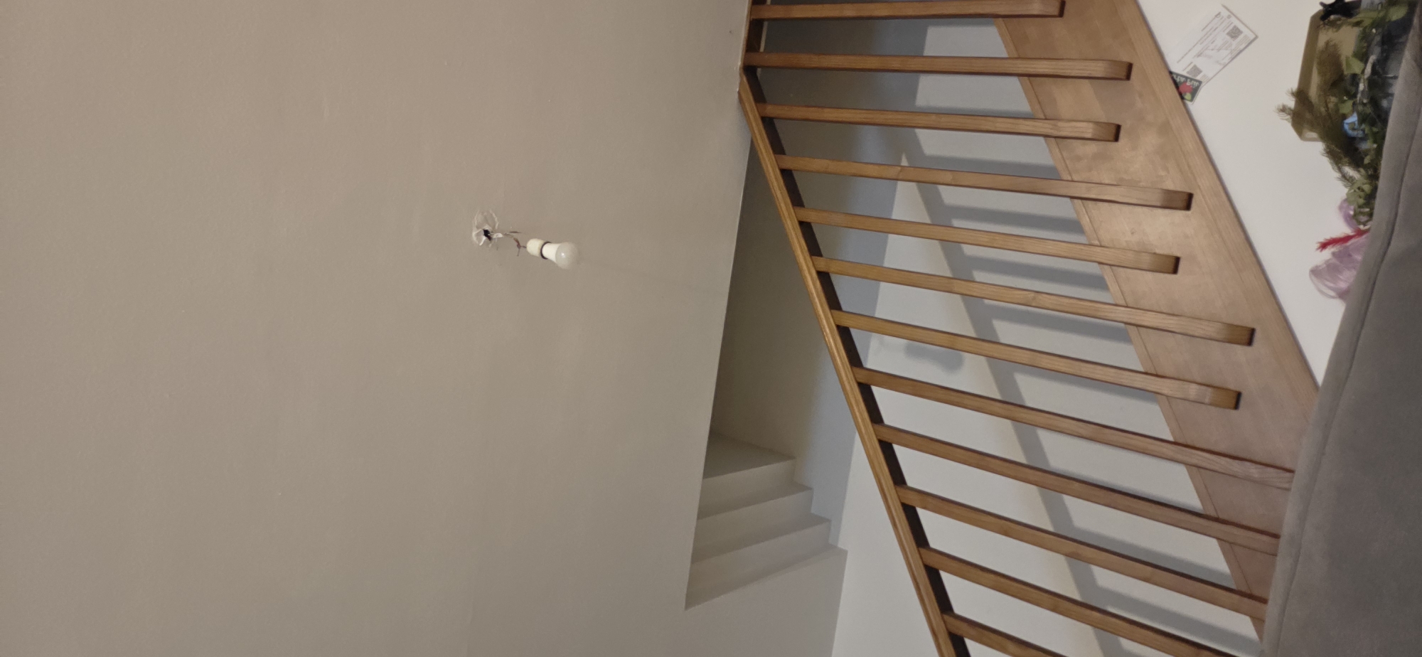 The image shows an interior scene with a wooden staircase and railing. Above the staircase, a bare lightbulb is hanging from the ceiling by its wires, which appear to be exposed. The ceiling is painted white. There is a recessed area with visible steps in the wall above the staircase. The railing is made of light-colored wood with vertical slats. Part of a gray sofa or couch is visible in the foreground at the bottom of the image, with some decorative items on it, including what appears to be a pink artificial flower and some greenery. The scene is devoid of any people. The image does not contain any information that would allow for its location to be identified as Farra di Soligo, Italy.
