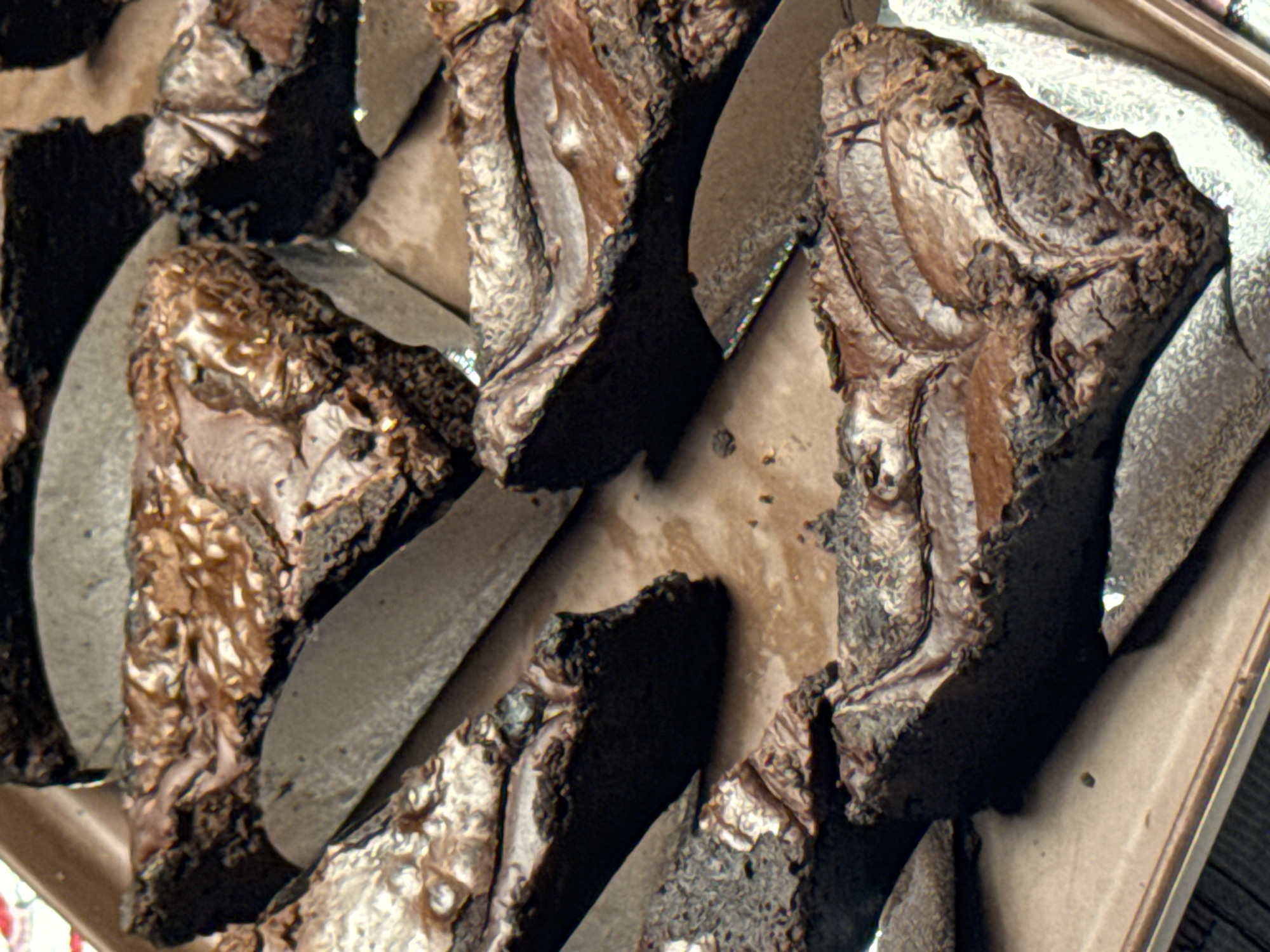The image displays a close-up, overhead view of a tray filled with freshly baked brownies. The brownies are cut into triangular slices and arranged in a slightly overlapping manner. They exhibit a rich, dark chocolate color with a glossy, swirled top. Some slices have a slightly cracked surface, characteristic of a well-baked brownie. The tray appears to be made of a light brown material, possibly cardboard or a similar disposable baking tray. The background is out of focus but suggests a light-colored surface, possibly a table or countertop. No people or specific landmarks are visible, making it impossible to determine the exact location in ตำบลเกาะพะงัน, Thailand, other than it is a food preparation or serving scene.