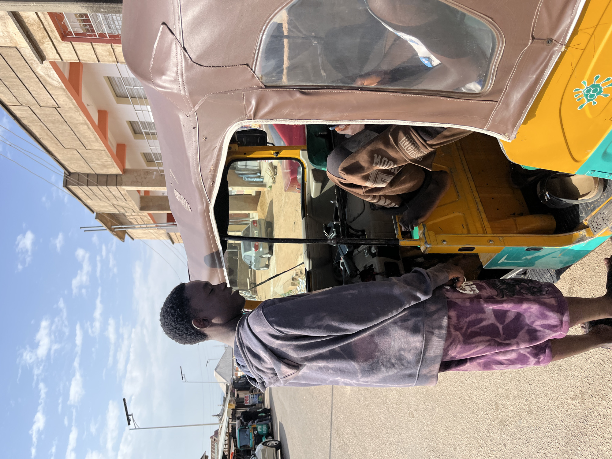 In Jos, Nigeria, a young man with short, dark hair stands beside a yellow and teal tuk-tuk on a paved street. He wears a gray, tie-dye sweatshirt and purple tie-dye shorts. The tuk-tuk has a brown canvas roof. A brown jacket rests on the back seat. Buildings line the street, and the sky is blue with scattered clouds. Other vehicles and pedestrians are visible in the background.