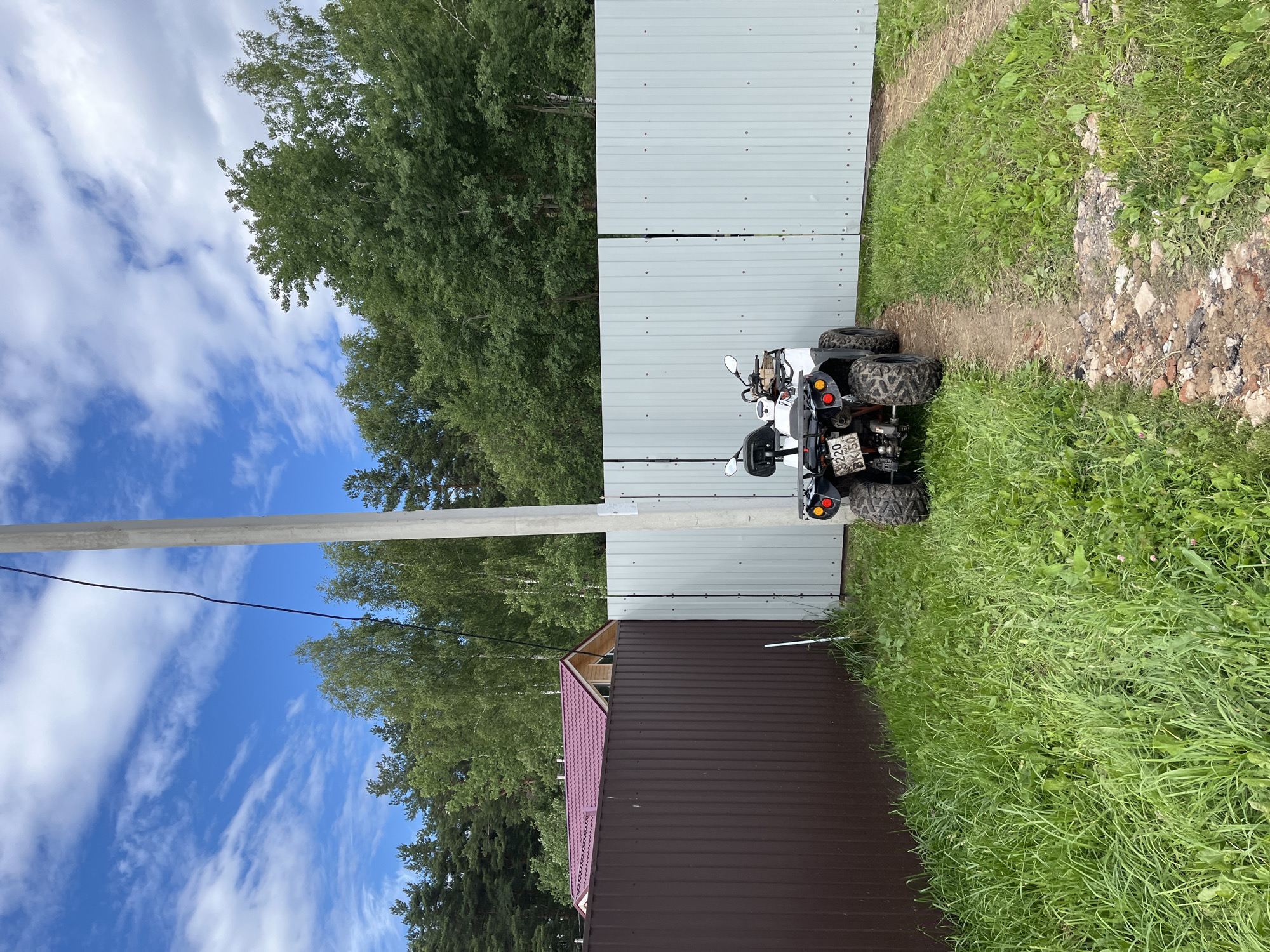 The media file depicts an outdoor scene in Razdolye, Russia. The main subjects are a white all-terrain vehicle (ATV) parked on a dirt path, a grey corrugated metal fence, a brown corrugated metal building with a reddish-purple roof, and lush green grass. In the background, there are numerous trees and a bright blue sky with wispy white clouds.
The ATV appears to be a recreational vehicle, likely parked for a break or storage. The fence and building suggest a rural or semi-rural residential setting, possibly a dacha or a farm. The time of day appears to be daytime, with the bright sky indicating fair weather. The overall atmosphere is peaceful and natural.
Notable details include the license plate on the ATV, which appears to be Russian, with Cyrillic characters. The details of the ATV include its headlights, handlebars, seat, and knobby tires. The building's roof has a distinctive corrugated pattern. The grass is long and appears somewhat unkempt, suggesting a natural or less manicured landscape.
There is no apparent activity or event taking place; the scene is static and appears to be a still photograph. The location cues of the ATV's license plate and the general landscape are consistent with a Russian setting. There are no visible emotions conveyed as there are no people in the image.