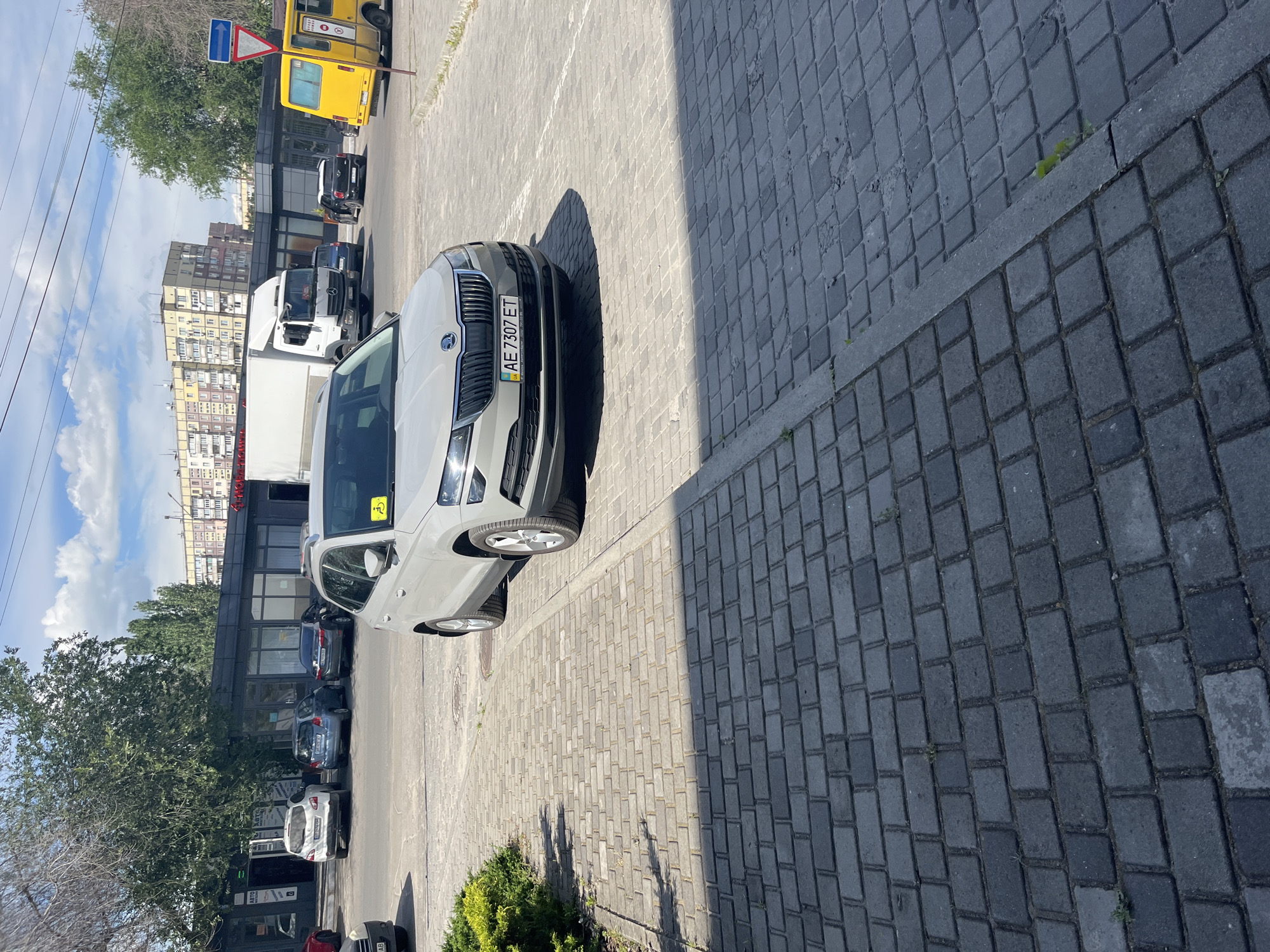 A light gray Skoda Karoq SUV is parked in a paved lot. The license plate reads AE 7307 ET. A yellow disability sticker is visible on the windshield. The car is facing forward, with its front end slightly angled towards the right. In the background, there is a low-rise building with large windows and an awning, and beyond that, taller apartment buildings. A white truck and a yellow bus are also visible in the background. The scene appears to be in an urban environment in Dnipro, Ukraine, as suggested by the car's license plate format and the architectural style of the buildings. The lot is paved with interlocking gray bricks, and there is a small green bush to the left of the car. The sky is blue with scattered white clouds. The lighting indicates it is daytime, likely late morning or early afternoon, with sunlight casting shadows.