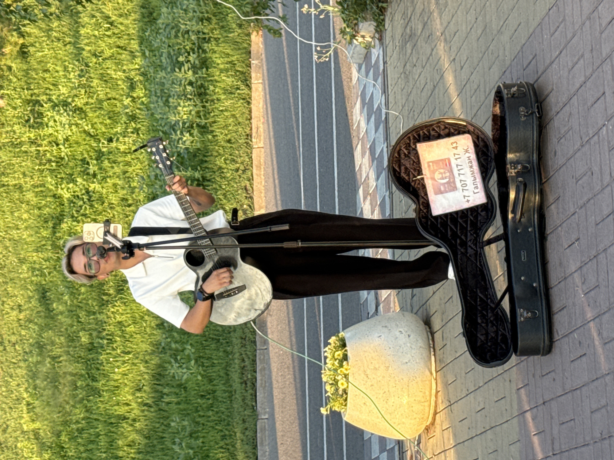 A male street performer is playing an acoustic guitar and singing into a microphone in Pavlodar, Kazakhstan. He is standing on a paved walkway next to a road, with a grassy embankment behind him. He is wearing a white zippered polo shirt and black pants, with light-colored hair and glasses. His guitar case is open on the ground in front of him, with a card inside that has a phone number and name. There is a large planter with yellow flowers to his left. The sun is casting long shadows, indicating it is late afternoon or early evening.