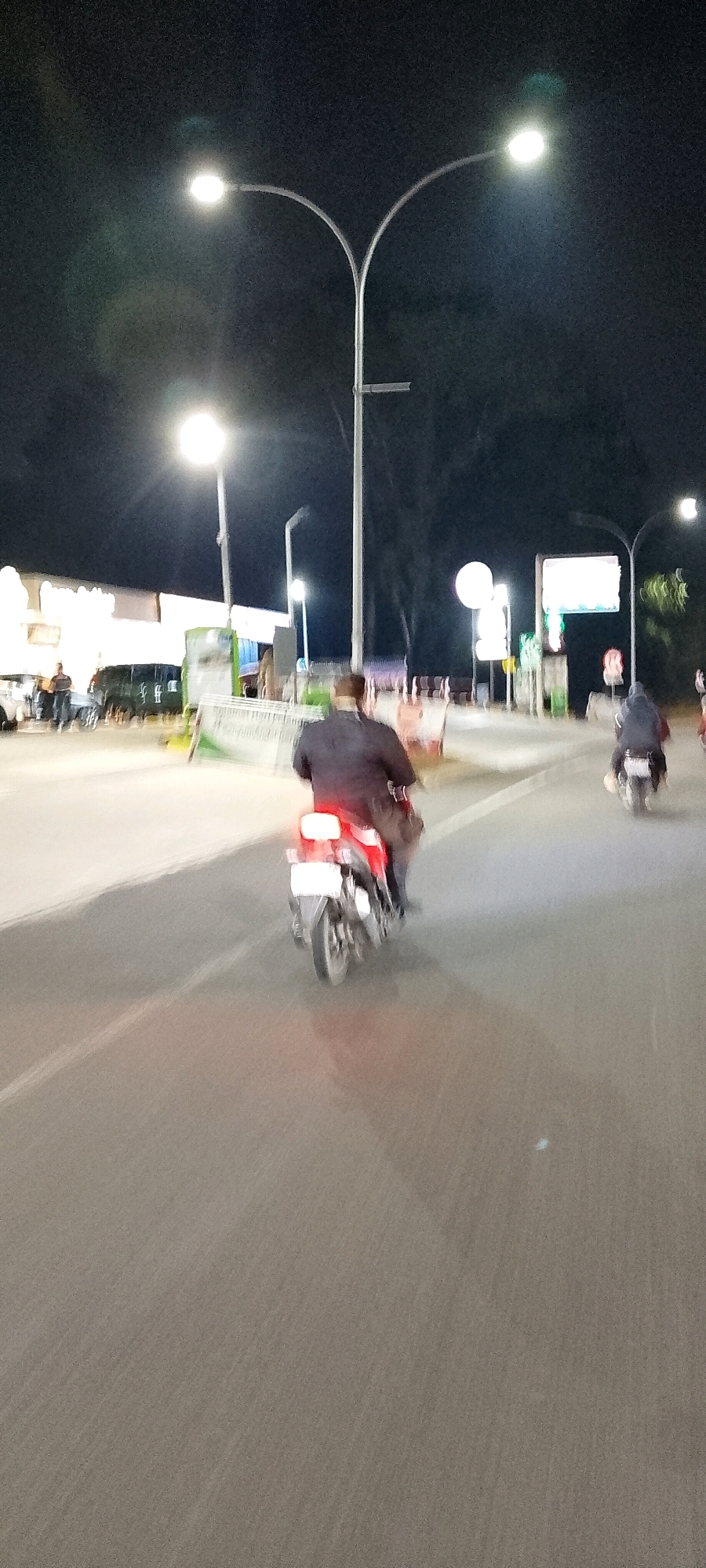 The image depicts a night scene on a road in Gunung Putri, Indonesia, illuminated by streetlights. A person is riding a motorcycle in the foreground, traveling away from the viewer. The motorcycle's brake lights are illuminated red. In the background, other vehicles and buildings are visible, though details are blurred due to motion and low light conditions. Two individuals are standing near a building on the left side of the image. Another motorcycle with a rider is visible further down the road on the right. The environment suggests a commercial or urban area with street lighting and structures.