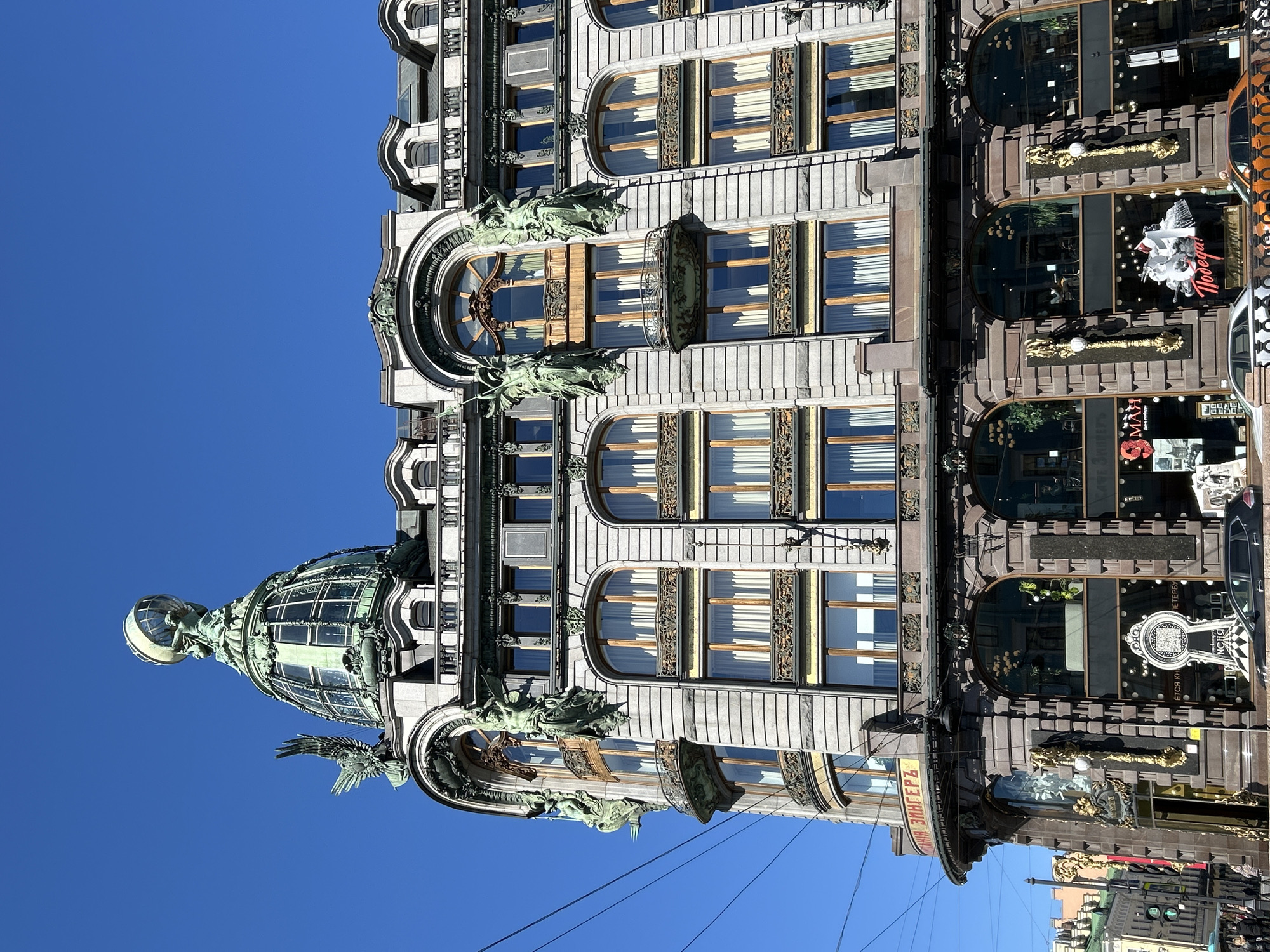 The image captures the Singer House in Saint Petersburg, Russia, viewed from a low angle. The architectural details of the building are prominently displayed, including its ornate facade, arched windows, and decorative sculptures. The sky is clear and blue. The lower part of the building features shop windows and the building's sign.