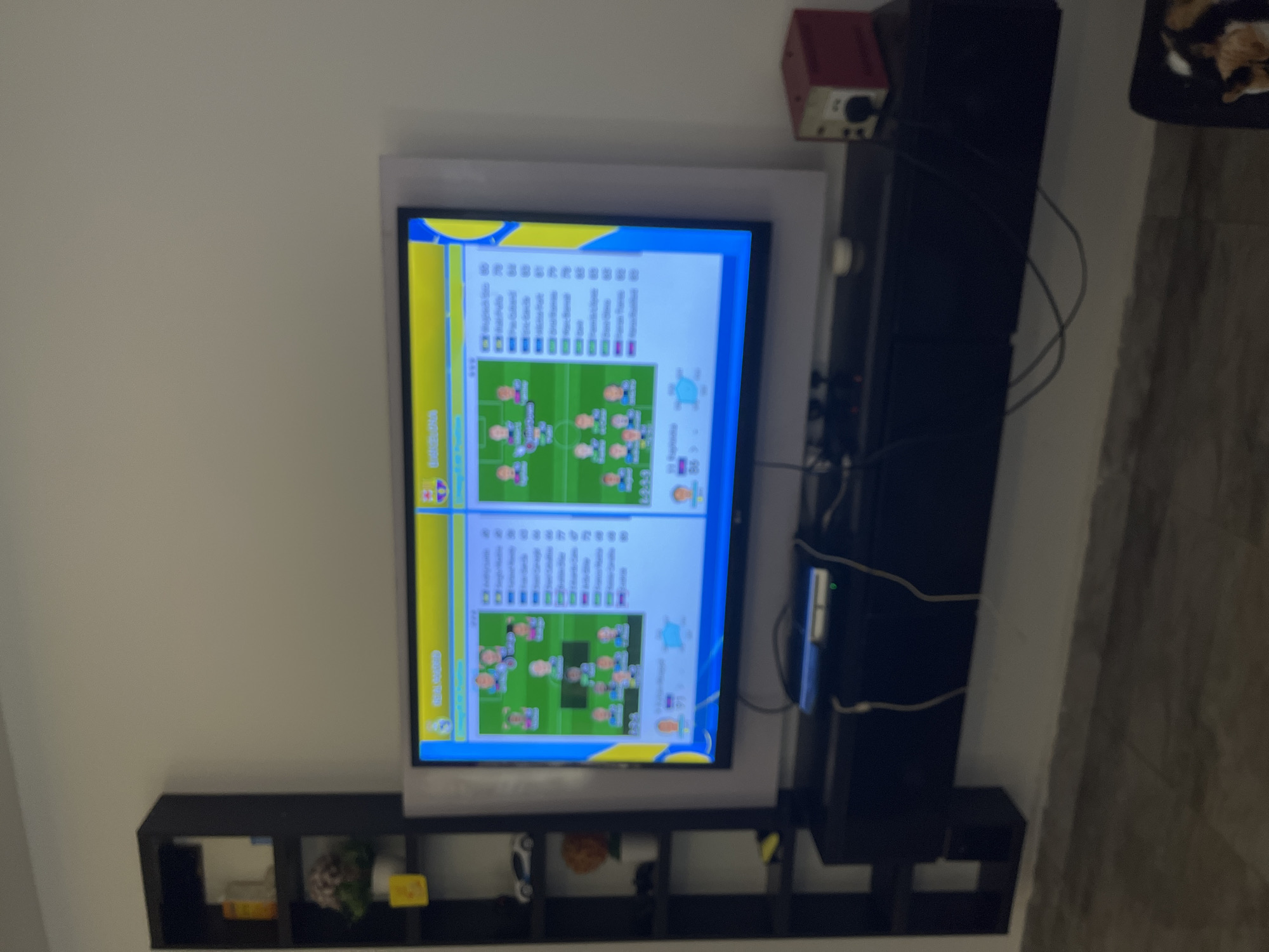 The image shows a television screen displaying a soccer team lineup. On the left side of the screen, the team "REAL MADRID" is shown with player icons and statistics. On the right side, the team "BARCELONA" is also displayed. The television is mounted above a black entertainment stand, which holds a game console and various cables. To the left of the television is a black, tall, narrow bookshelf with several shelves, containing decorative items, a toy car, and a potted plant. The scene appears to be indoors, with a light-colored wall and tiled flooring visible. The lighting is moderate, suggesting it is daytime or the room is well-lit. The overall environment seems to be a living room or entertainment area. The image does not provide any information about the city of Nabatum, Nigeria.