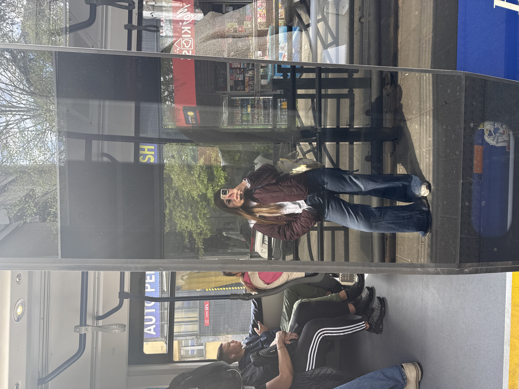 The image depicts the interior of a public transportation vehicle, specifically a bus, with passengers seated. The scene suggests the location is Vozdovac Urban Municipality, Serbia based on the reflection visible through the bus window.
Several passengers are present. One individual, possibly a young woman, is reflected in the bus window while holding a phone. Other passengers, including a male with a backpack, a person wearing Adidas-striped pants, and others in casual clothing, are seated.
The environment includes elements typical of a bus interior, such as handrails and advertisements visible on the window frame. Reflections outside the bus window include a building with signage and general urban scenery. The bus seems to be in motion.