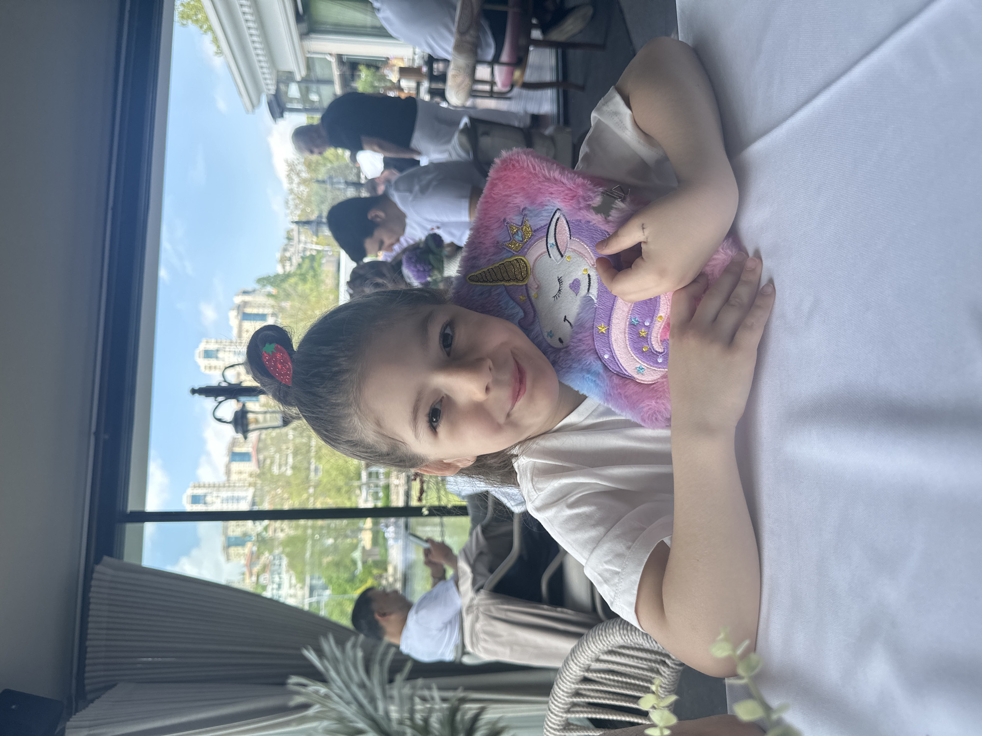 A young girl with strawberry-shaped hair clips is holding a fluffy unicorn notebook and smiling at the camera. She is sitting at a table with a white tablecloth. Through a large window, a cityscape is visible, suggesting a location in Başakşehir, Turkey. Other people are seated at tables in the background, indicating a restaurant or cafe setting.
