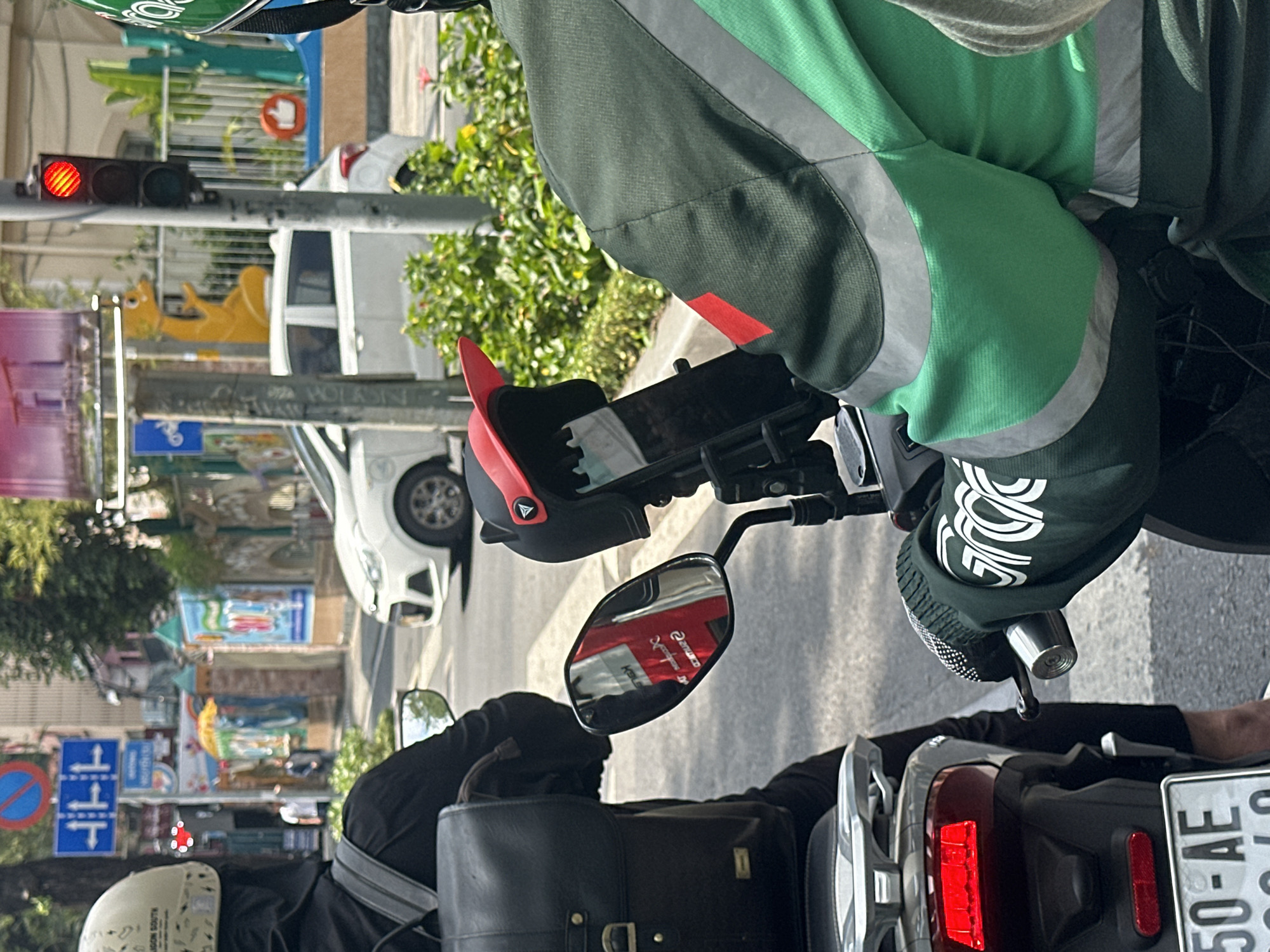 A street scene in Thủ Đức, Vietnam, depicts two individuals on scooters halted at a red traffic light. In the immediate foreground on the right, a person is partially visible, wearing a dark green, light green, and grey jacket with reflective strips. The person's left arm, clad in a black sleeve with white "GRAB" branding, grips a scooter handlebar. A black helmet with a red visor attachment is mounted on the scooter's front, holding a mobile phone. Ahead, to the left, a second individual is seen from behind, wearing a black long-sleeved garment and a black head covering under a light-colored helmet inscribed with "SAIGON SOUTH." This person is seated on a light grey scooter, which displays a red taillight and a partial Vietnamese license plate "50-AE". A black leather-like backpack is worn. The scooter's rearview mirror reflects a red and white sign. In the background, a white Hyundai i10 sedan is stationary on the asphalt road, which features a visible crosswalk. A red traffic light is illuminated on a pole. Multiple blue road signs are visible, one indicating lane directions. Green bushes line the sidewalk. Building facades are visible beyond, featuring colorful murals, including a prominent yellow sculptural figure resembling a creature and a red "thumbs up" icon on a blue background.