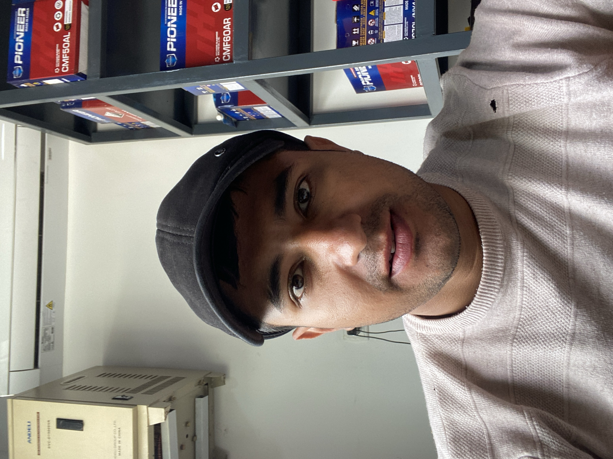 A close-up portrait of a man wearing a black cap and a light pink sweater, situated indoors. Behind him, a metal shelf holds stacks of "PIONEER" branded boxes, some labeled "CMF50AL" and "CMF60AR", suggesting these are car batteries. A cream-colored electrical device with the brand "ANDELI" is also visible, bearing the text "MADE IN CHINA." The location and the city of Bukhara, Uzbekistan cannot be verified from the visual information.