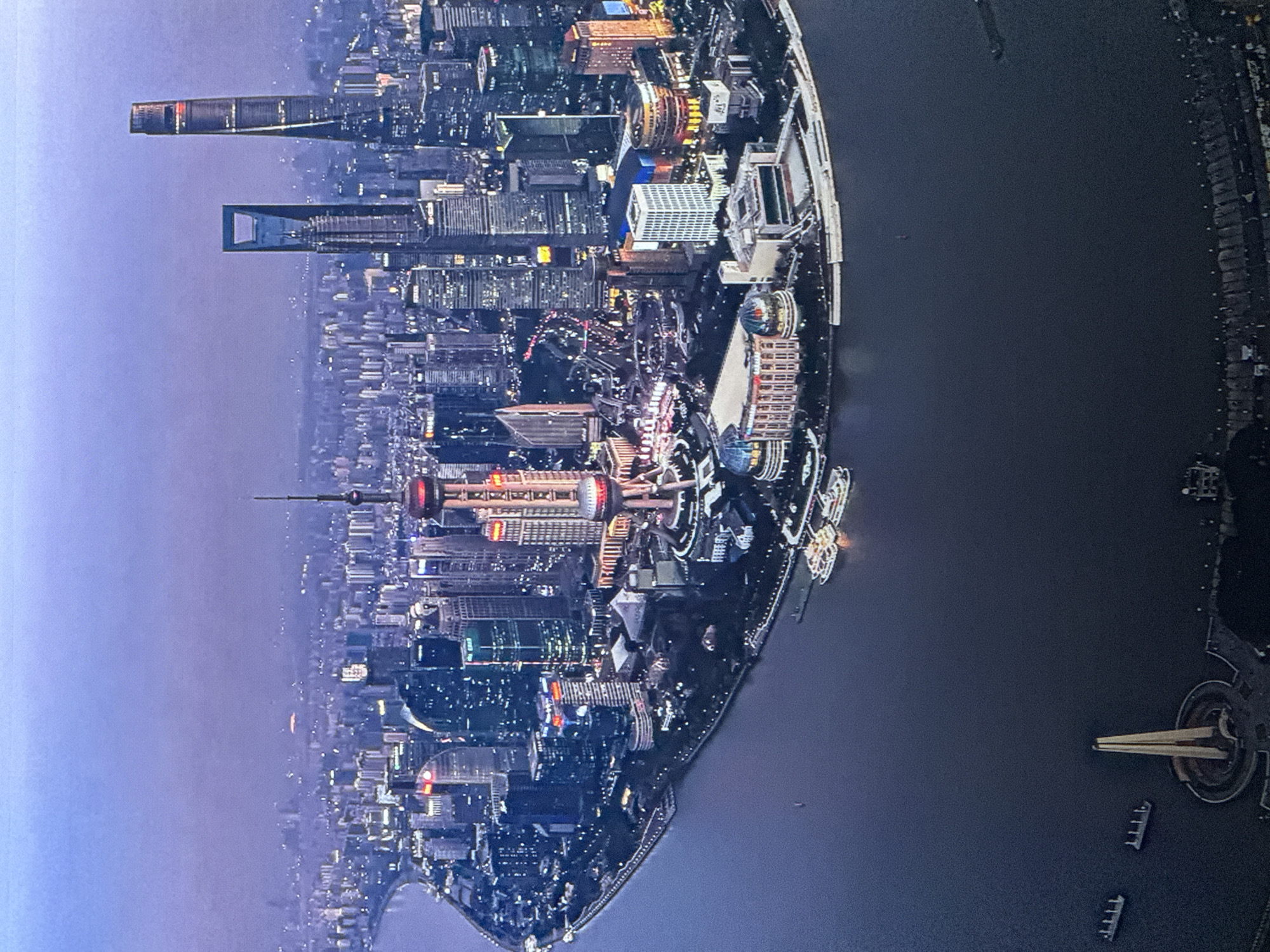 The image is an aerial view of the Shanghai skyline at dusk. The city is illuminated by numerous lights from skyscrapers and other buildings. In the foreground, a wide body of water, likely the Huangpu River, dominates the lower half of the frame. Several boats, including some larger passenger ferries docked along the shore, are visible on the water. The cityscape features a dense collection of high-rise buildings, with distinct landmarks like the Oriental Pearl Tower and the Shanghai Tower. The sky is a hazy twilight blue, suggesting either pollution or the transition from day to night. There is no indication of Saratov, Russia in the image.