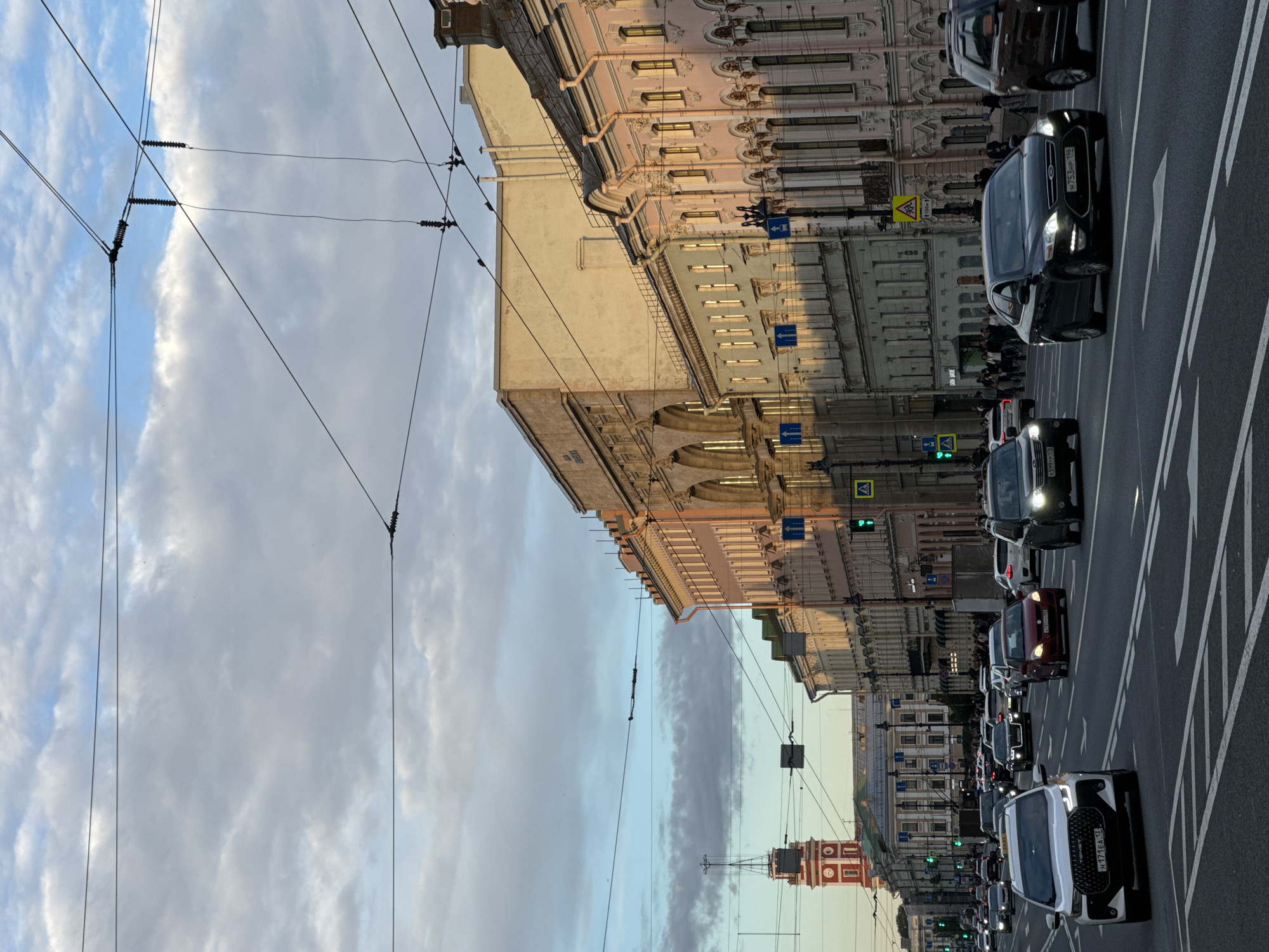 The image depicts a wide street in Saint Petersburg, Russia, during daylight. The sky is partly cloudy, with soft sunlight illuminating the buildings. The street is busy with traffic, including cars, vans, and a bus. Pedestrians are visible on the sidewalks, and traffic lights and street signs are present. The architecture of the buildings lining the street is varied, with some featuring classical elements and others being more modern. In the background, a red tower with a clock face stands out against the skyline. The overall impression is one of a vibrant urban environment on a pleasant day.