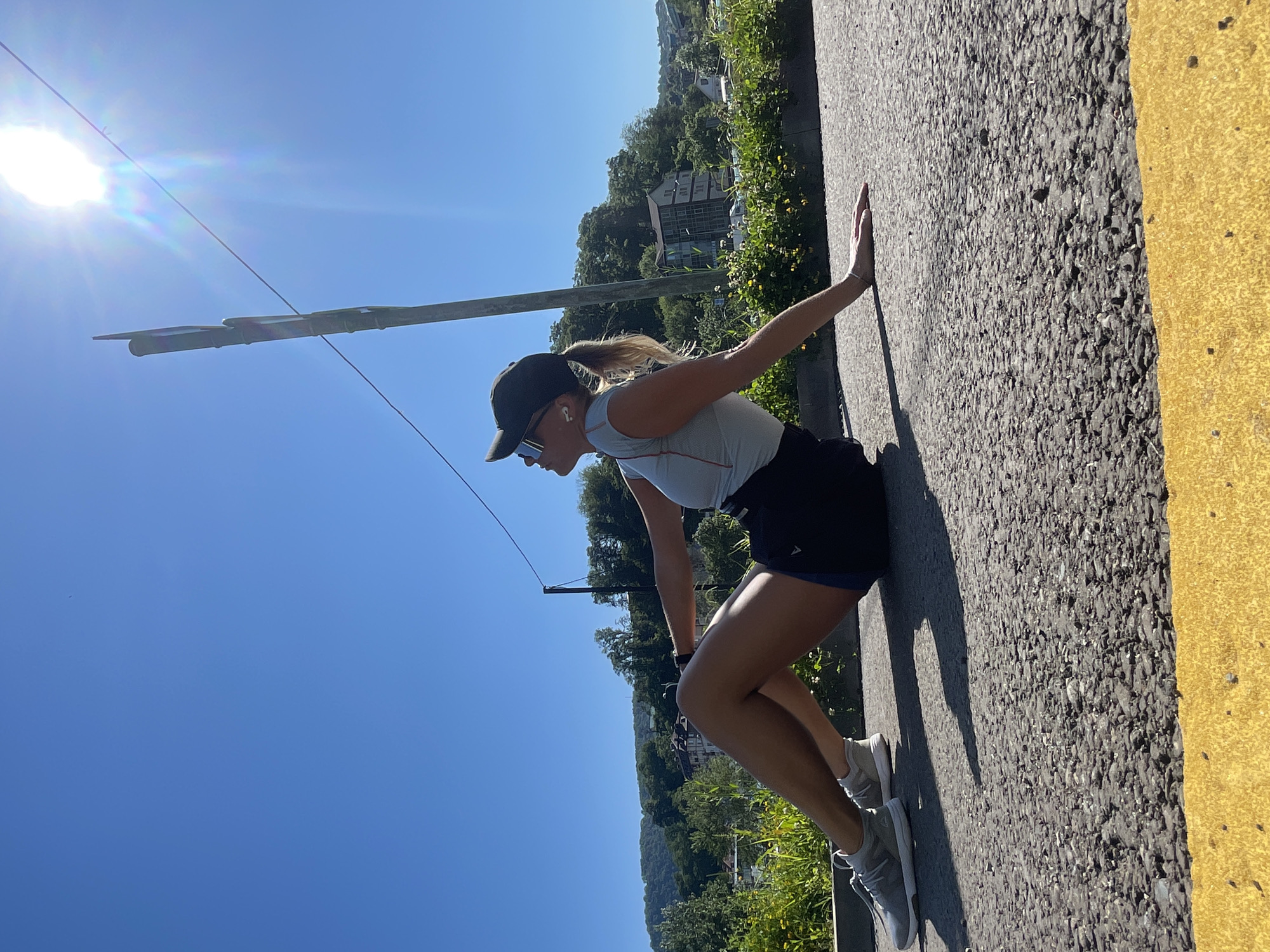 A woman is captured in a dynamic pose outdoors on a bright, sunny day, likely in Sochi, Russia. She is leaning forward with her hands pressed against a textured asphalt surface, seemingly performing a wall-push-up, stretch, or resistance exercise, with her body angled almost parallel to the surface. She appears focused on her activity.
The woman is dressed in athletic attire, including a dark baseball cap, mirrored sunglasses, a light grey or white sleeveless athletic top with a subtle red stripe, dark athletic shorts with blue trim, and light grey athletic shoes. Her hair, pulled back in a ponytail, is visible beneath her cap, and she wears white wireless earbuds. A smartwatch is on her left wrist.
The scene is set under a clear, vibrant blue sky with the sun shining brightly, causing a lens flare in the upper left corner. Behind the woman, a verdant hillside rises, densely covered with trees and lush green foliage. Several buildings, possibly residential structures, are visible nestled within the greenery on the hillside. A utility pole or similar horizontal structure cuts across the sky in the middle ground. The asphalt surface she is interacting with features a distinct yellow painted line, suggesting a road or path. The overall impression is one of active outdoor recreation in a scenic, warm environment. There is no discernible text visible on her clothing or in the immediate surroundings.