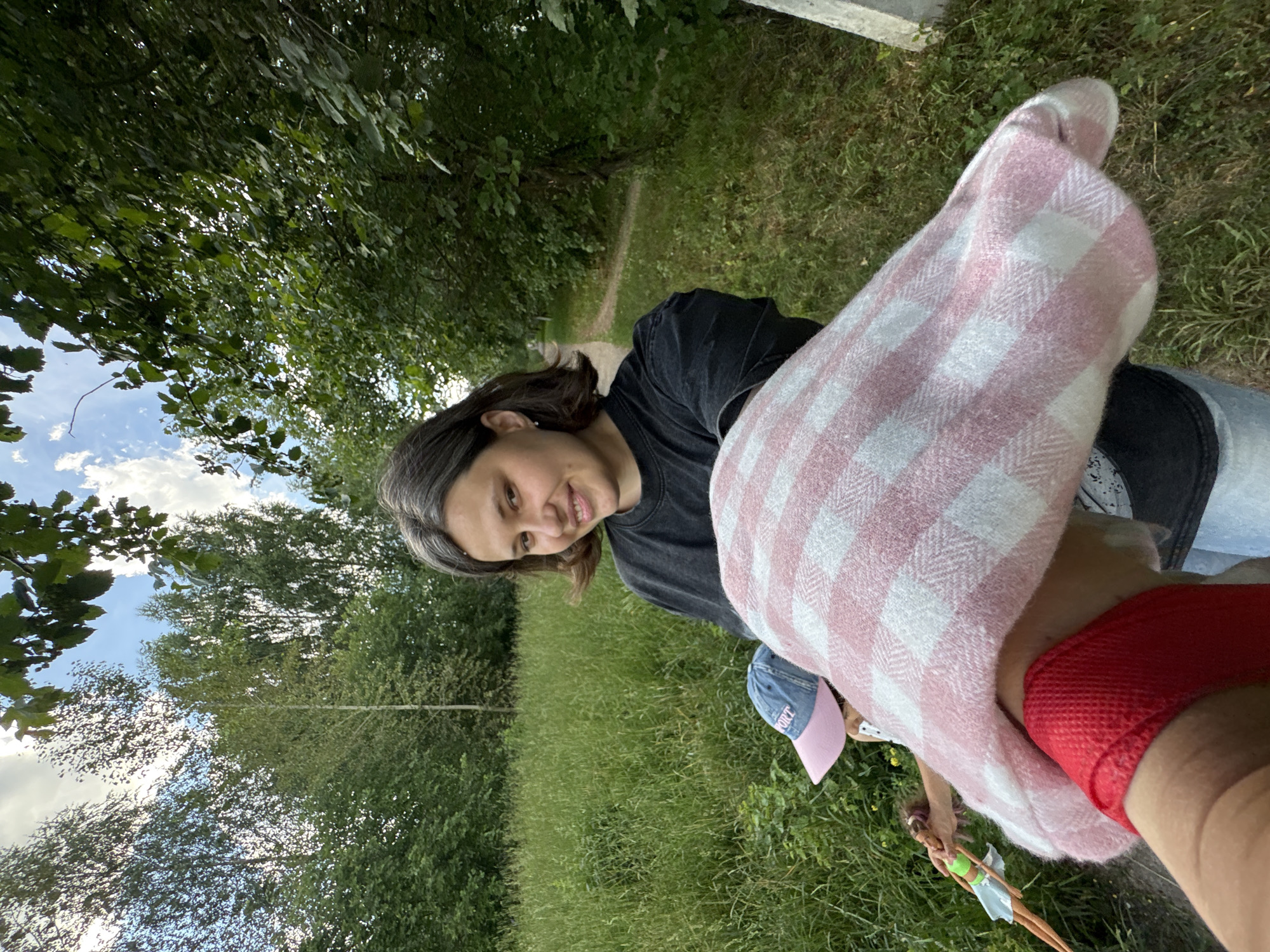 A young woman with short, graying dark hair and a black t-shirt smiles at the camera. She is in an outdoor setting, with tall green grass and trees visible in the background. A path winds through the grass behind her. A child wearing a pink and blue baseball cap is partially visible in front of her, holding a doll. The woman's arm is extended forward, holding a pink and white checkered blanket or fabric. The image appears to be taken in a park or a natural area within the city of Chaikovsky, Russia.