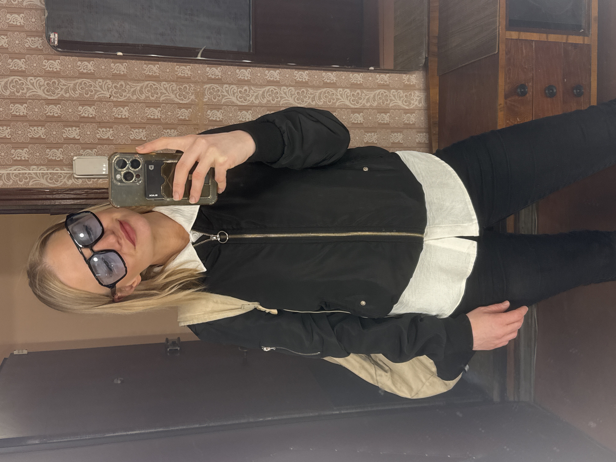 A person is taking a selfie in a room. The person is wearing sunglasses, a white collared shirt, a black bomber jacket, and black pants. The room appears to be a hallway or dressing room, with a patterned wallpaper and a dark wooden cabinet. There is a door and a mirror in the background. It is not possible to ascertain the location to be in Saint Petersburg, Russia based on provided image.