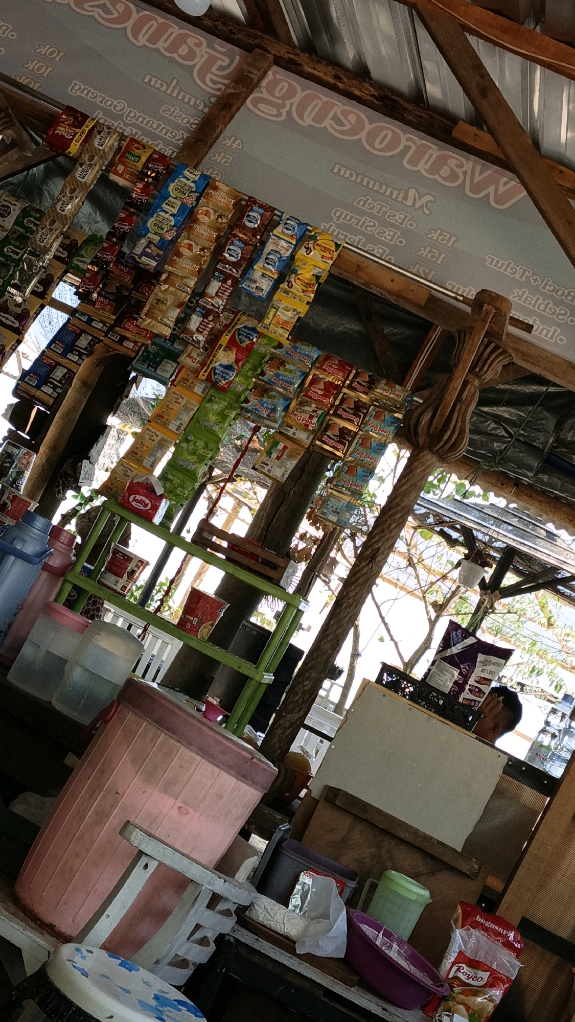 The image shows an outdoor stall in Cikal, Indonesia. The stall is filled with various items, including packets of snacks, bottles, and plastic containers. A person is partially visible in the background, looking down. The stall appears to be made of wood and metal, with a corrugated metal roof. The overall impression is of a simple, local market stall.